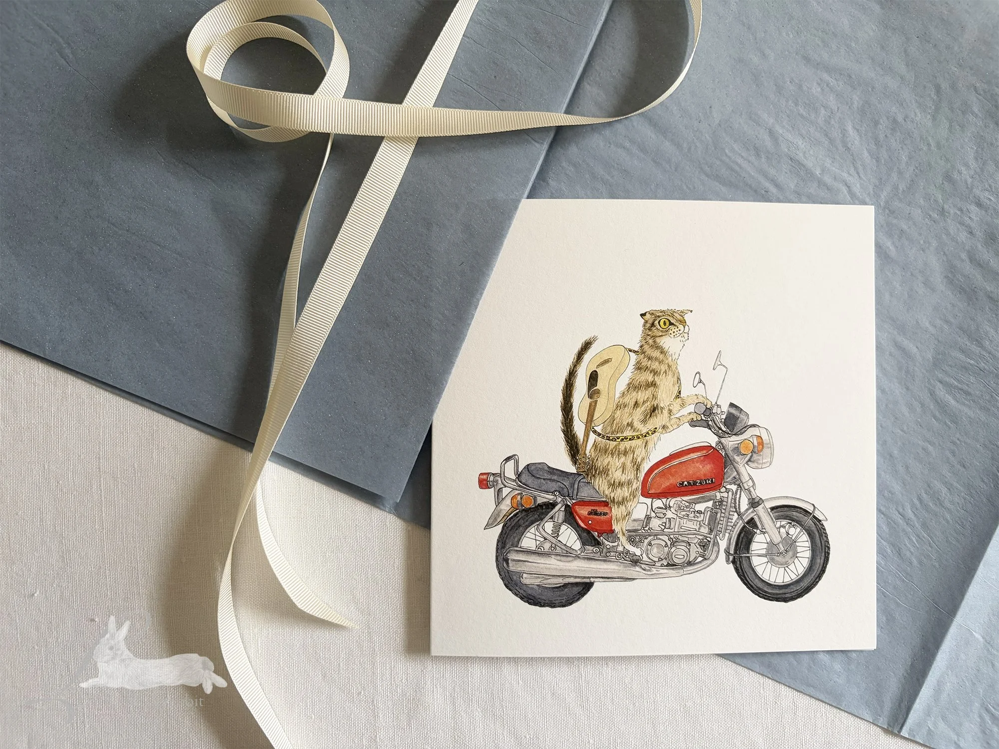 Hand painted greetings card showing a tabby cat on a red motorbike with a guitar on his back, sitting on a white linen tablecloth with cream ribbon, blue tissue paper, and a wax seal with stamp.