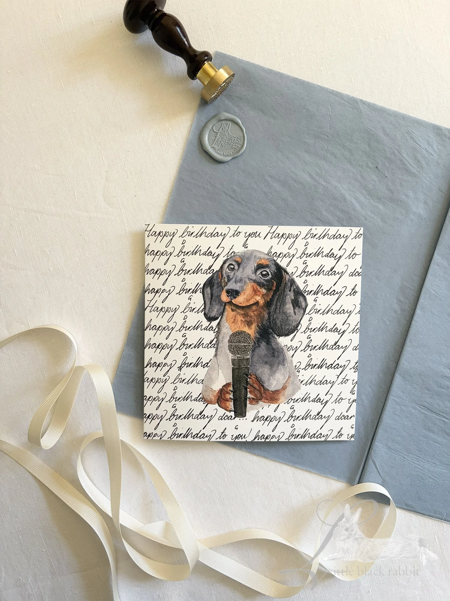 Sausage dog on a birthday card featuring cursive 'Happy Birthday' writing in the background, sitting on a table with a white linen tablecloth and dusty blue tissue paper.