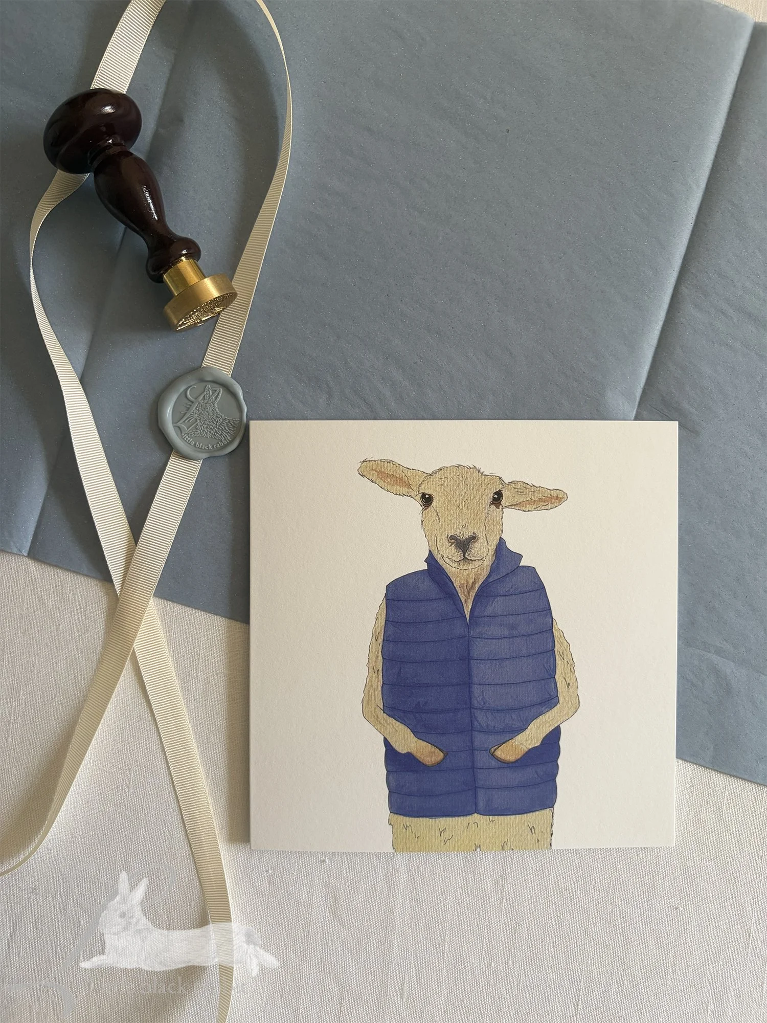 Handpainted greeting card featuring a smiling sheep in a blue quilted gilet, sitting on a white linen tablecloth with blue tissue paper, cream ribbon, and a blue wax seal.