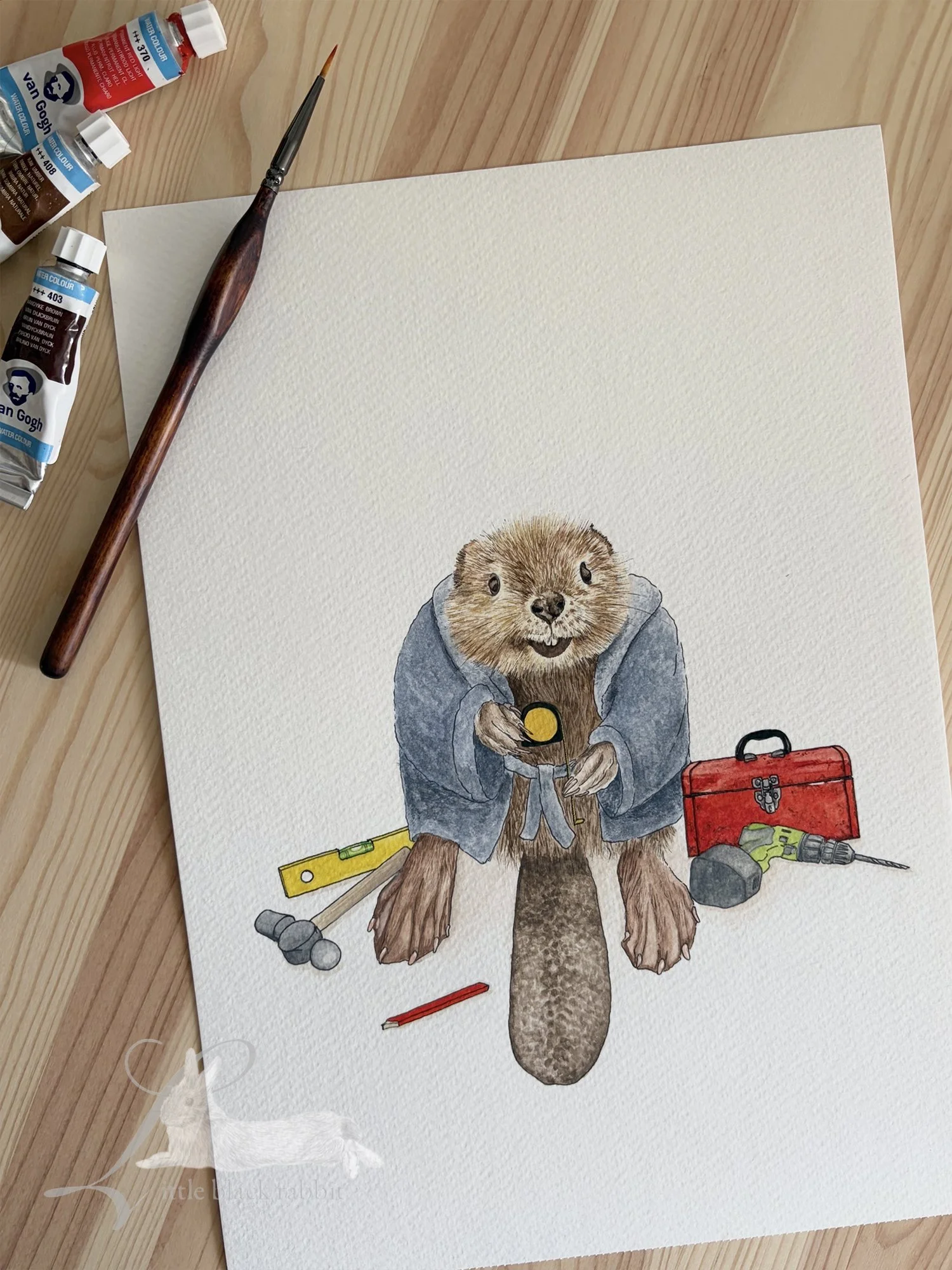 Original watercolour painting of a beaver wearing a blue dressing gown and sitting amongst a selection of tools and a tool box, sitting on a wooden table with paintbrushes and pencils.