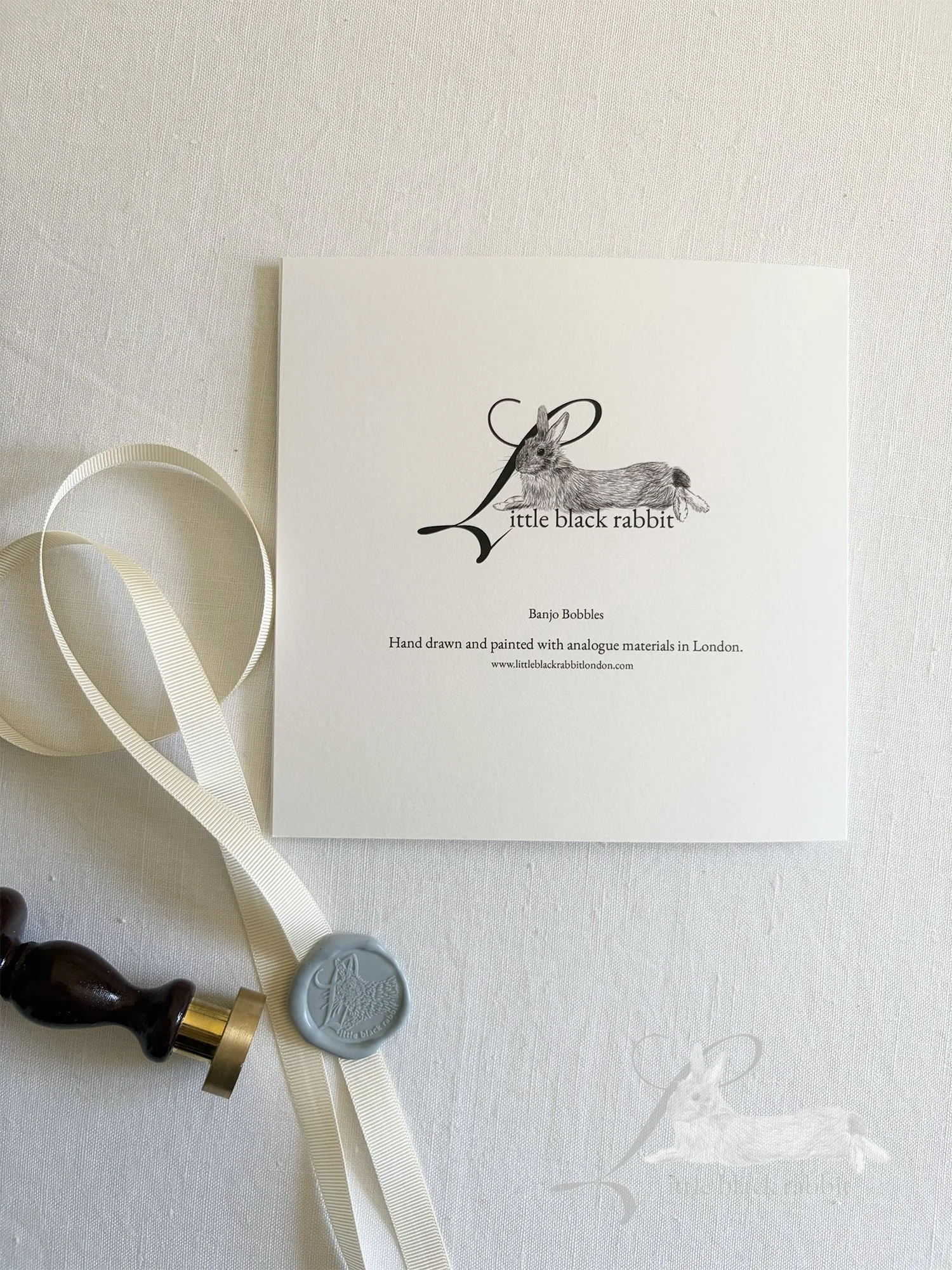 Reverse side of a hand painted greeting card showing the Little Black Rabbit logo, sitting on a white linen tablecloth with blue tissue paper, cream ribbon and a wax seal.