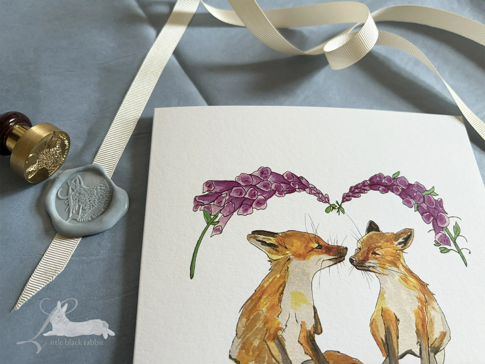 Close up image of a greetings card depicting two fox cubs nose to nose under some foxgloves, on a table with a wax seal and somce cream ribbon.