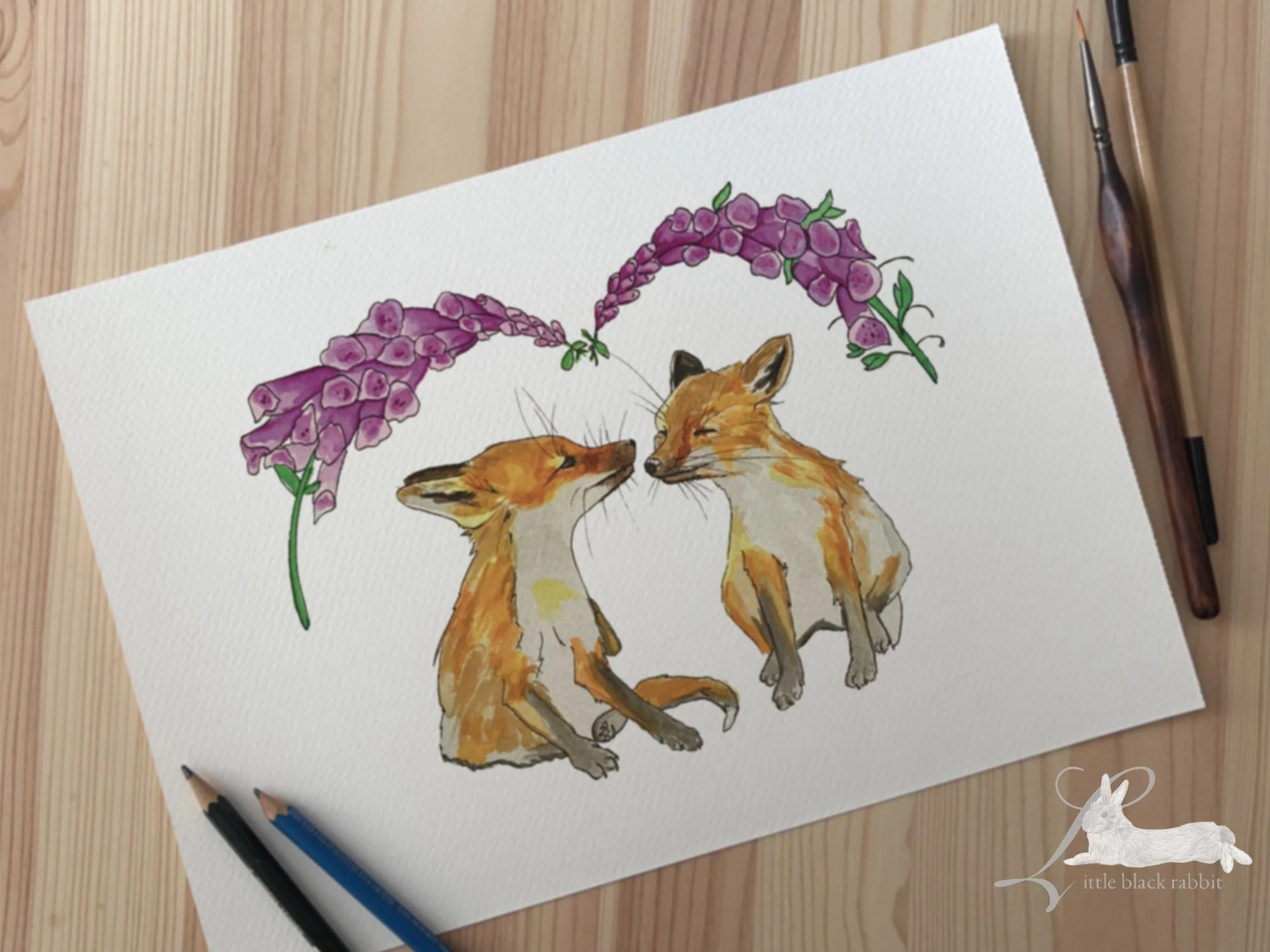Original watercolour illustration of a pair of fox cubs sitting nose to nose under some pink foxgloves, with a paintbrush on a wooden table.