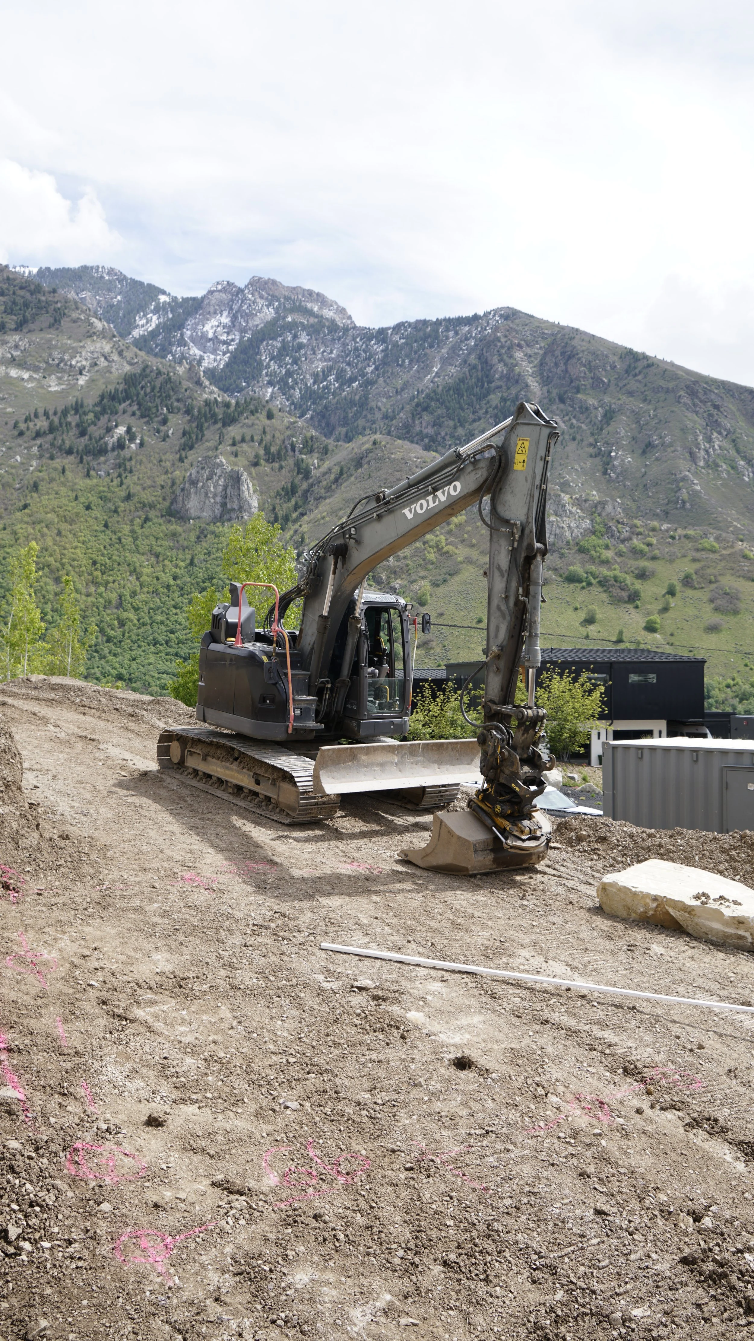 A construction site with a small Volvo excavator on dirt, with mountains in the background and pink chalk markings on the ground.