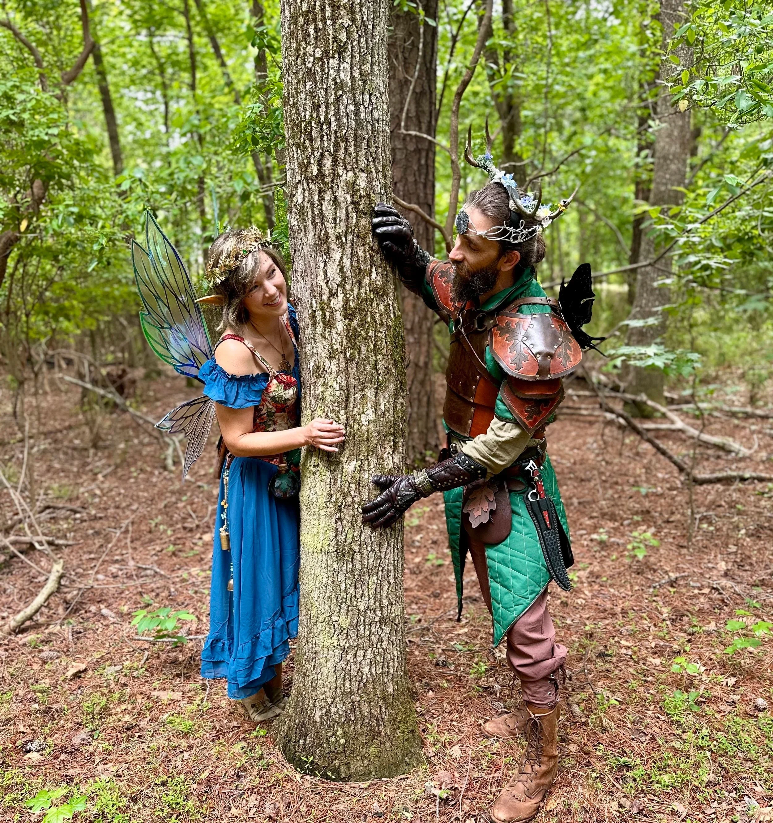 Two people in fairy and elf costumes with wings and antler crowns, sharing a moment by a tree in a forest, dressed in fantasy and medieval-inspired attire.