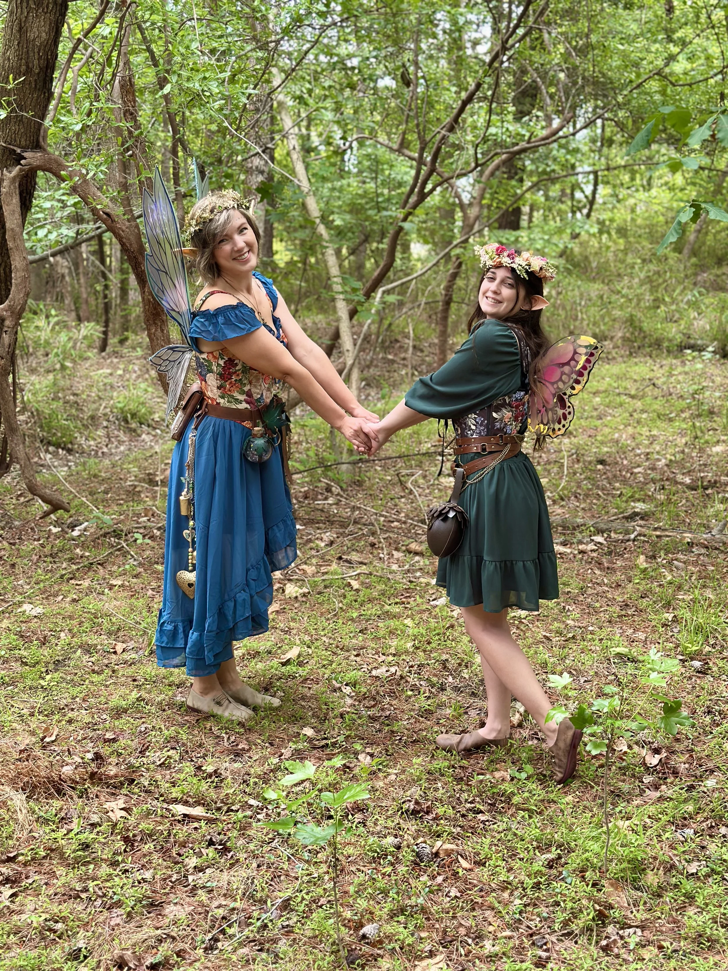 Two women dressed as fairies holding hands in a wooded forest, wearing fairy wings, floral crowns, and flowy dresses, with natural greenery and pinecones on the ground.