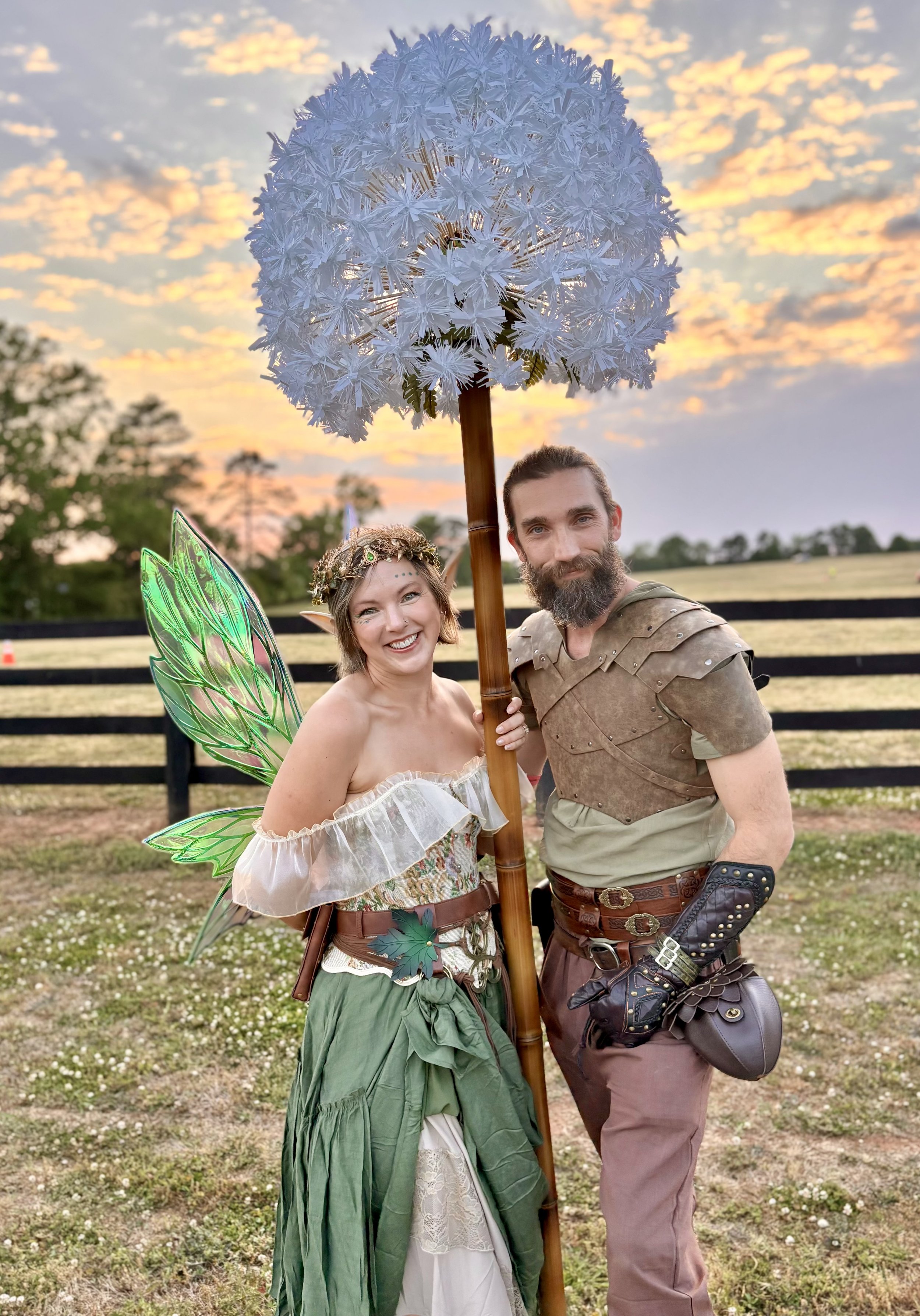 Two people dressed as fantasy characters, a fairy and a knight, standing outdoors at sunset. The woman has fairy wings, a crown, and a long green skirt. The man has a beard, medieval-style clothing, and armor accessories. They are smiling and holding a large, decorative umbrella resembling a cluster of white flowers.