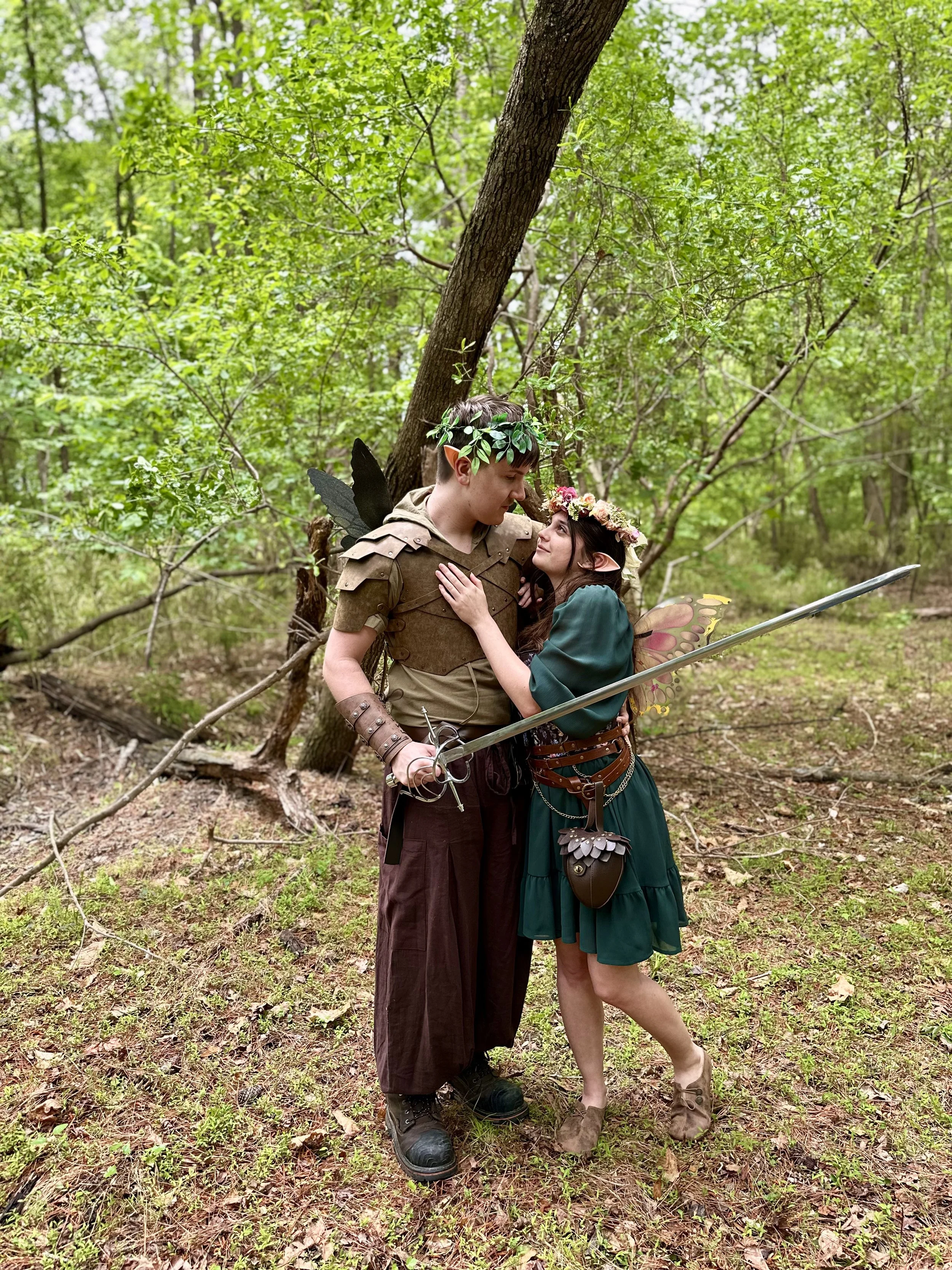 A fantasy-themed couple dressed as a fairy and an elf in a lush green forest, posing closely together with the fairy holding a sword.