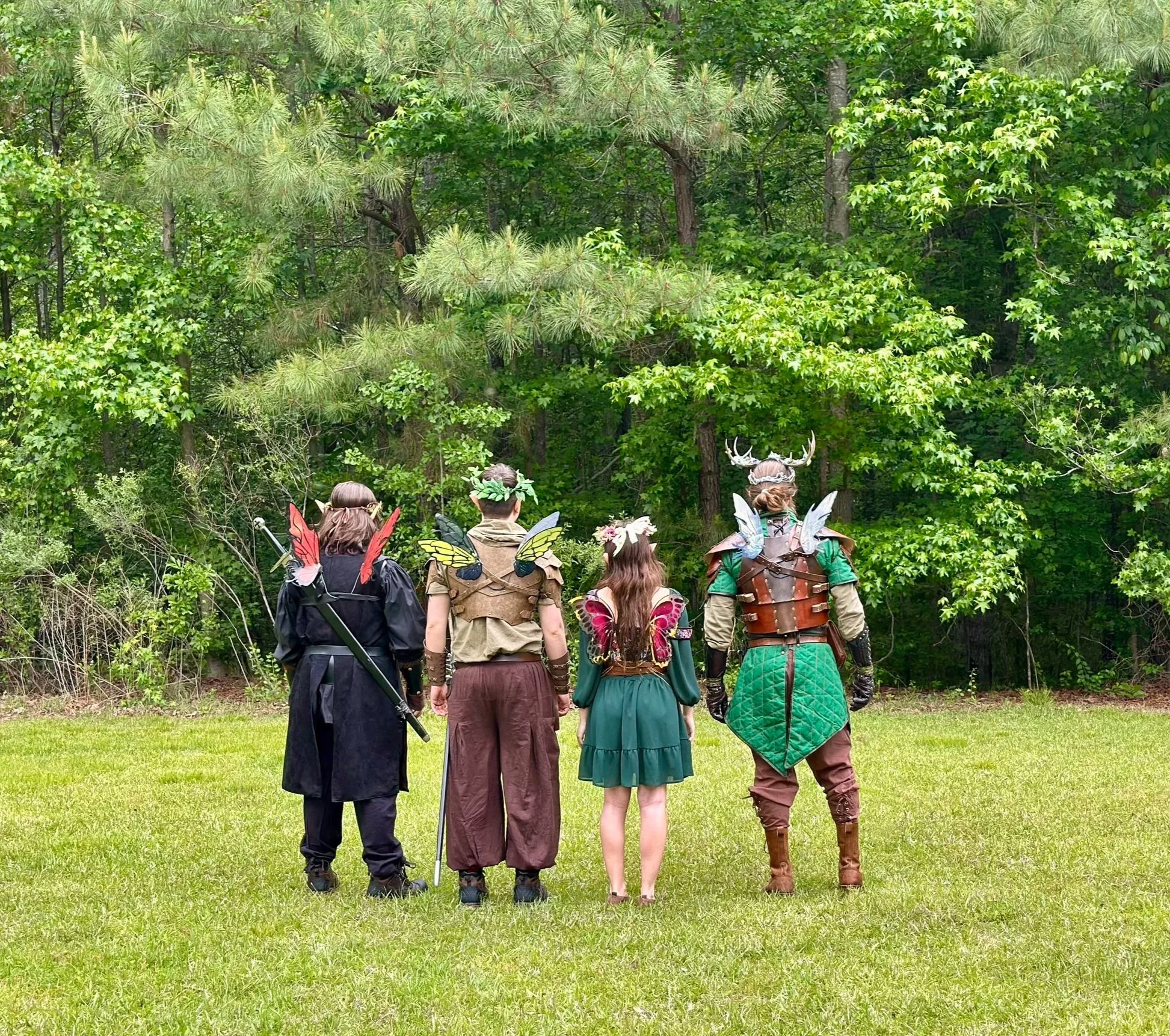 Group of people dressed as fantasy characters with wings, costumes, and accessories, standing on grass near trees.
