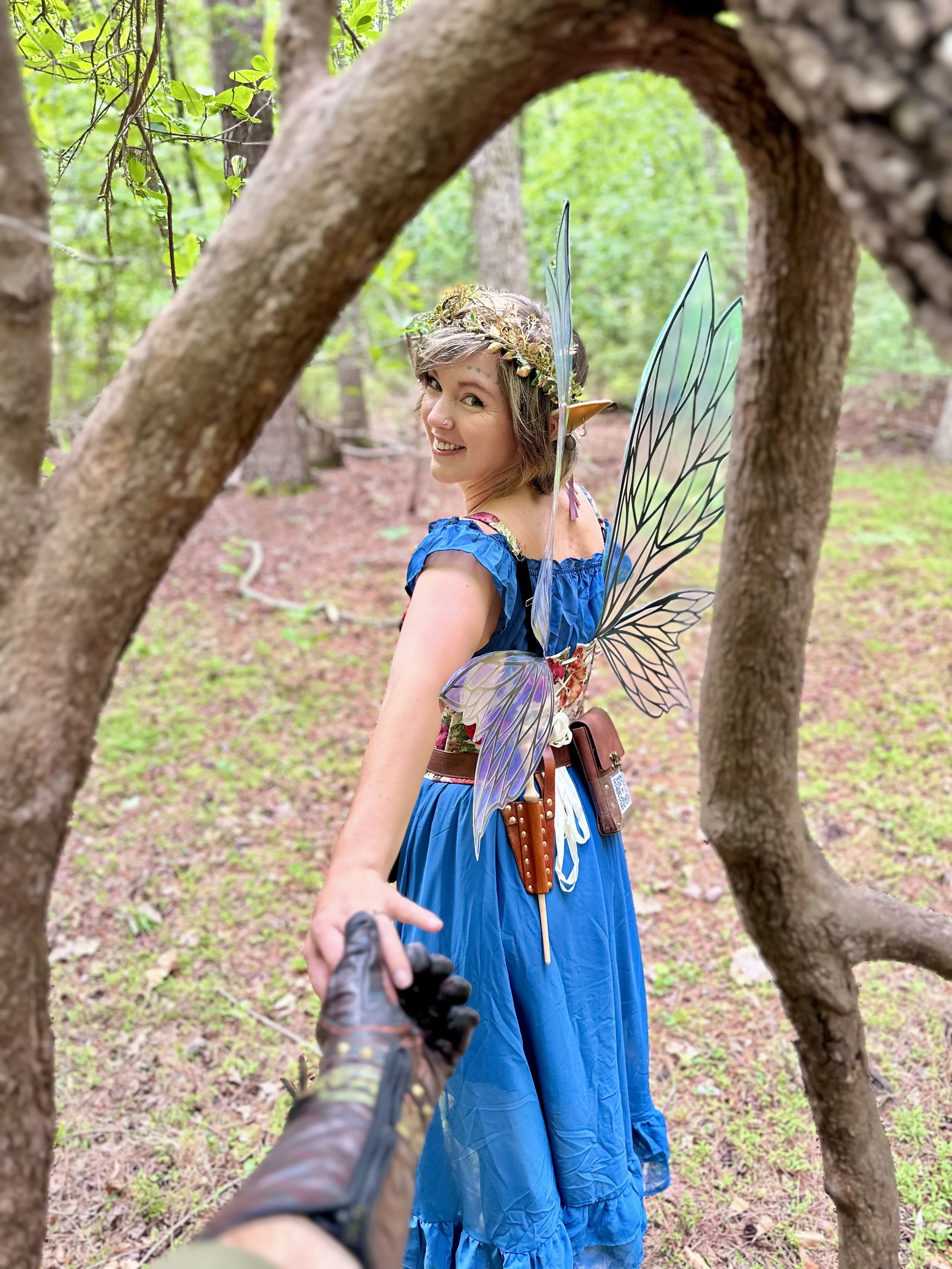 A woman dressed as a fairy with blue dress, butterfly wings, a floral crown, and elf ears, smiling and looking back in a wooded forest, holding hands with someone else.