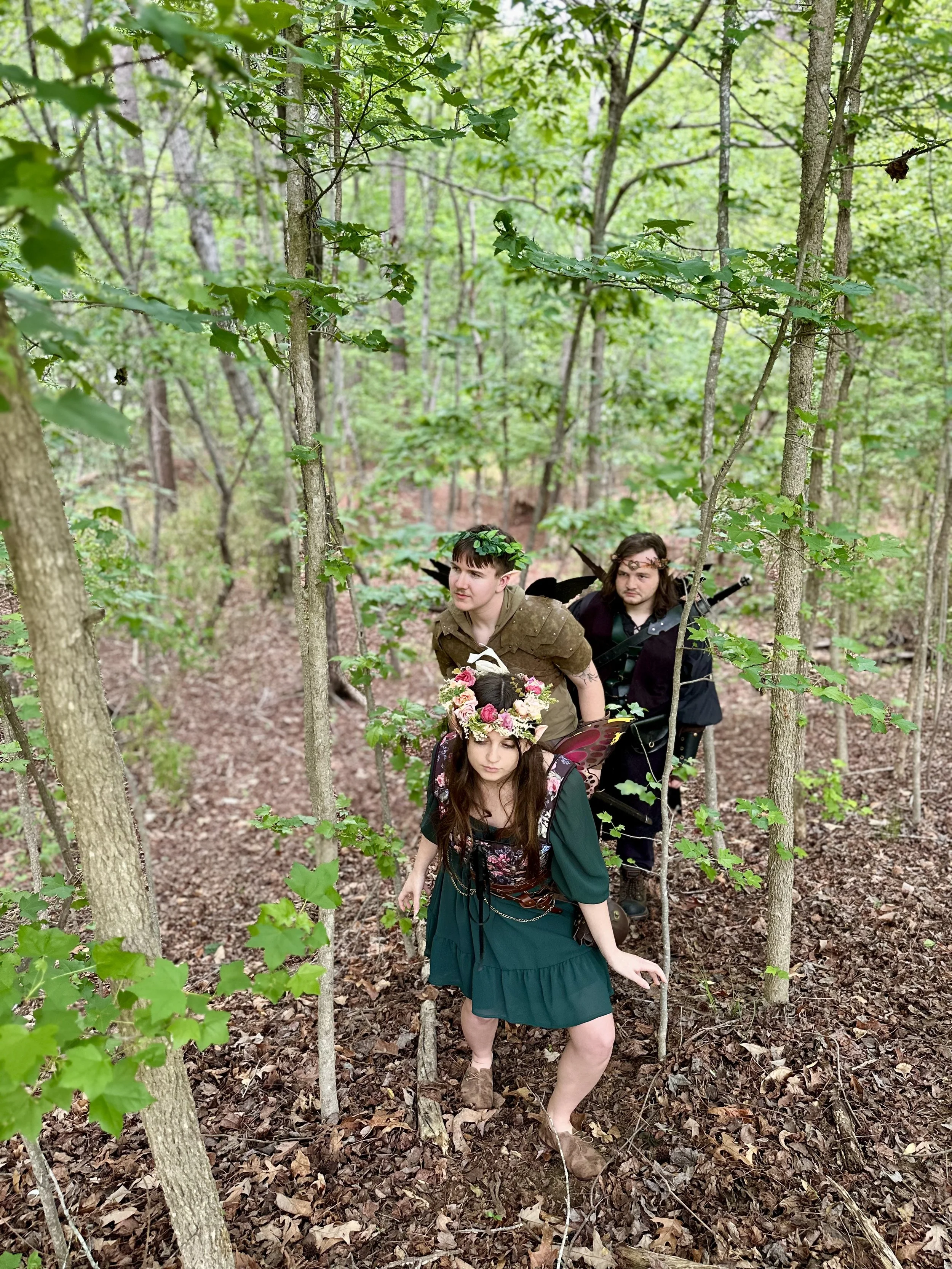 Three people dressed as fantasy characters, possibly fairies or elves, in a densely wooded forest, with one person in front wearing a floral crown and green dress, and two behind wearing dark clothing with wings.