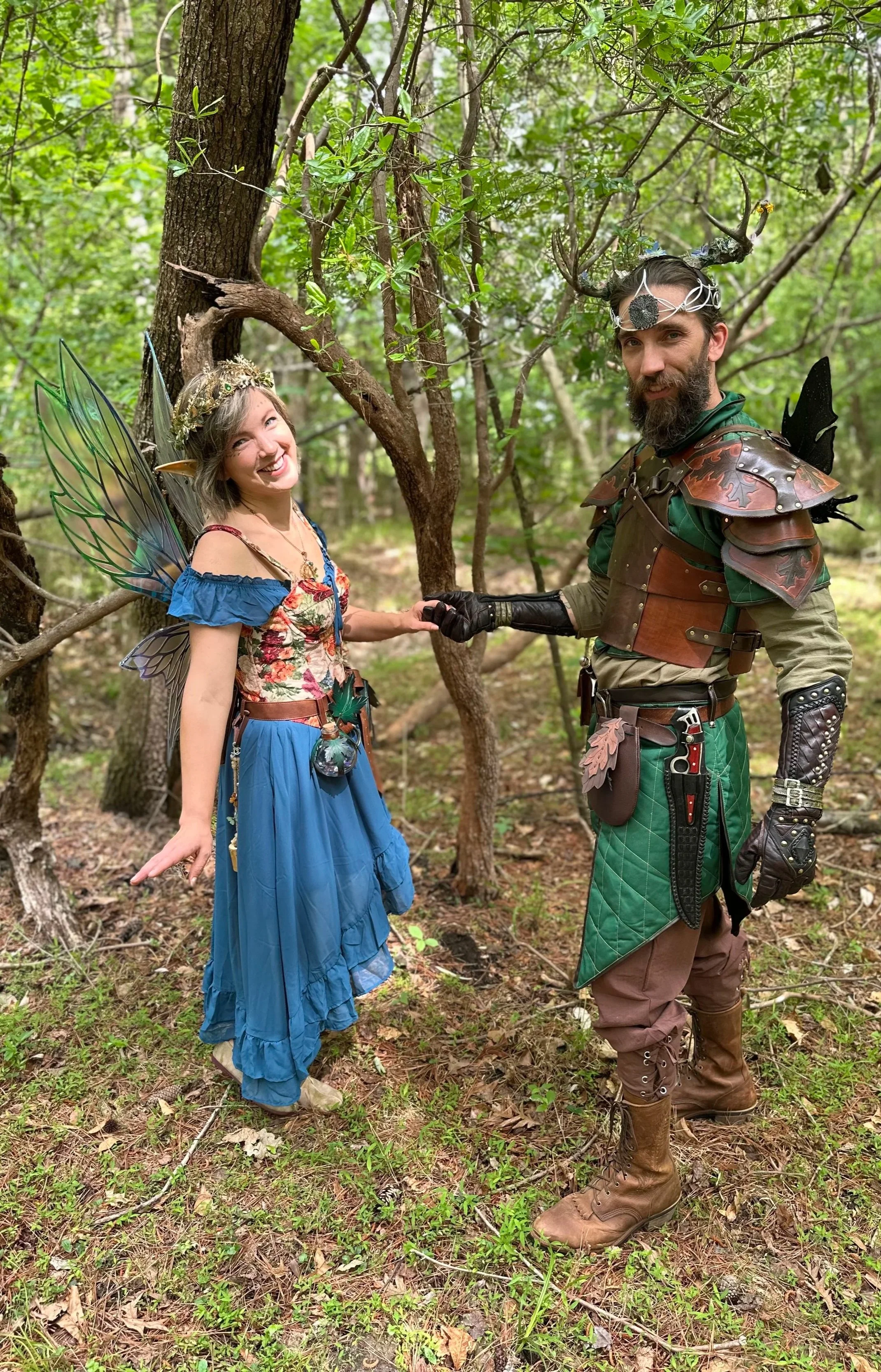 A woman dressed as a fairy with wings and a blue dress shaking hands with a man dressed as a fantasy warrior in a green and brown costume with armor and antler headpiece, in a forest with trees and green foliage.
