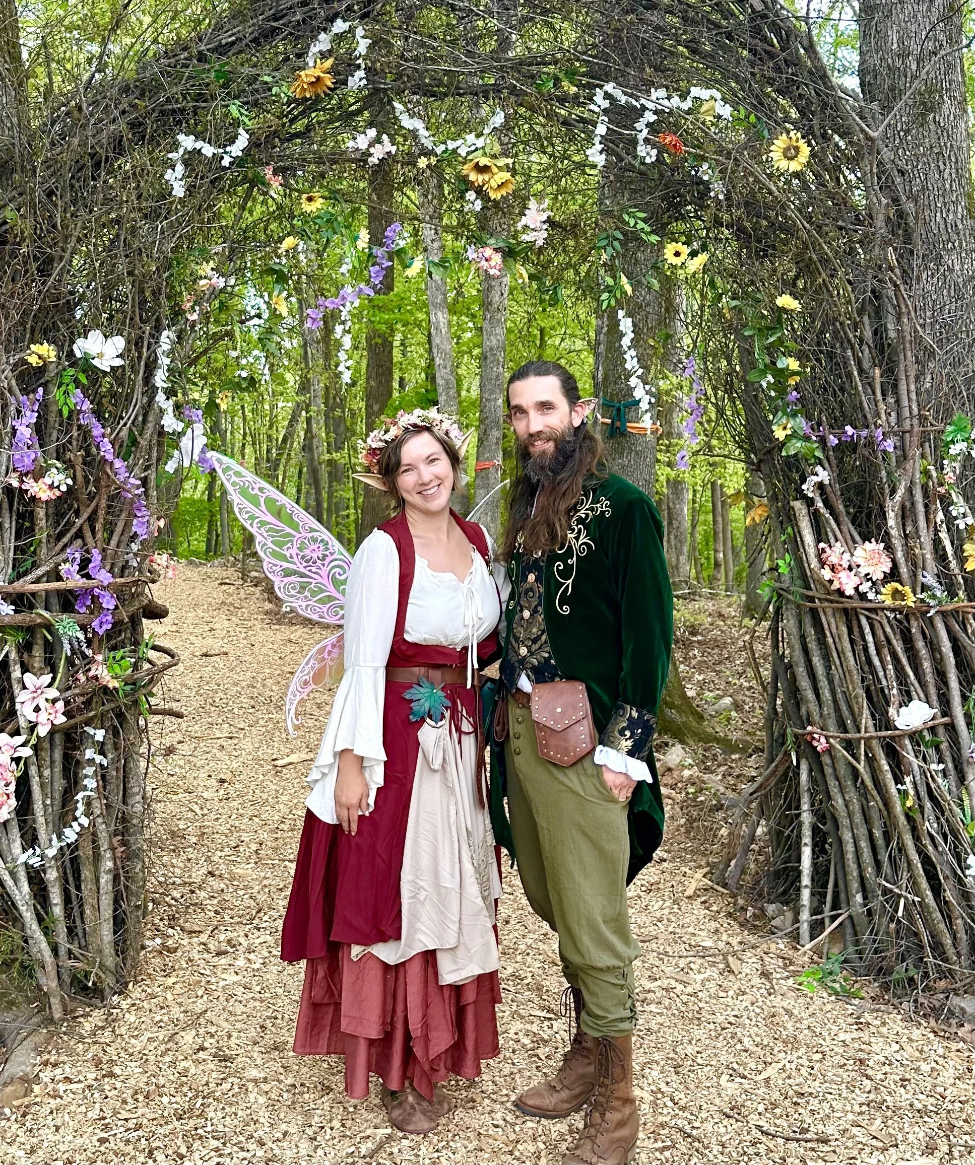 Two people dressed as fantasy characters standing in front of a wooden arch decorated with flowers and greenery in a forest setting.