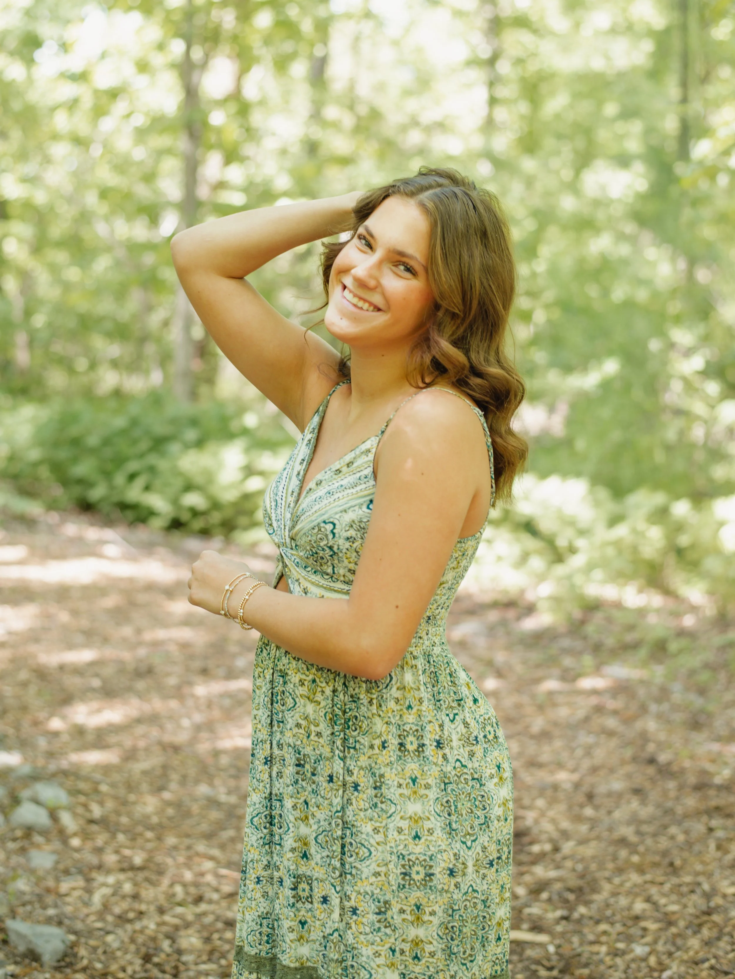 A young woman with wavy brown hair smiling outdoors in a wooded area, wearing a sleeveless yellow dress with a blue and green pattern, and bracelets on her wrist.