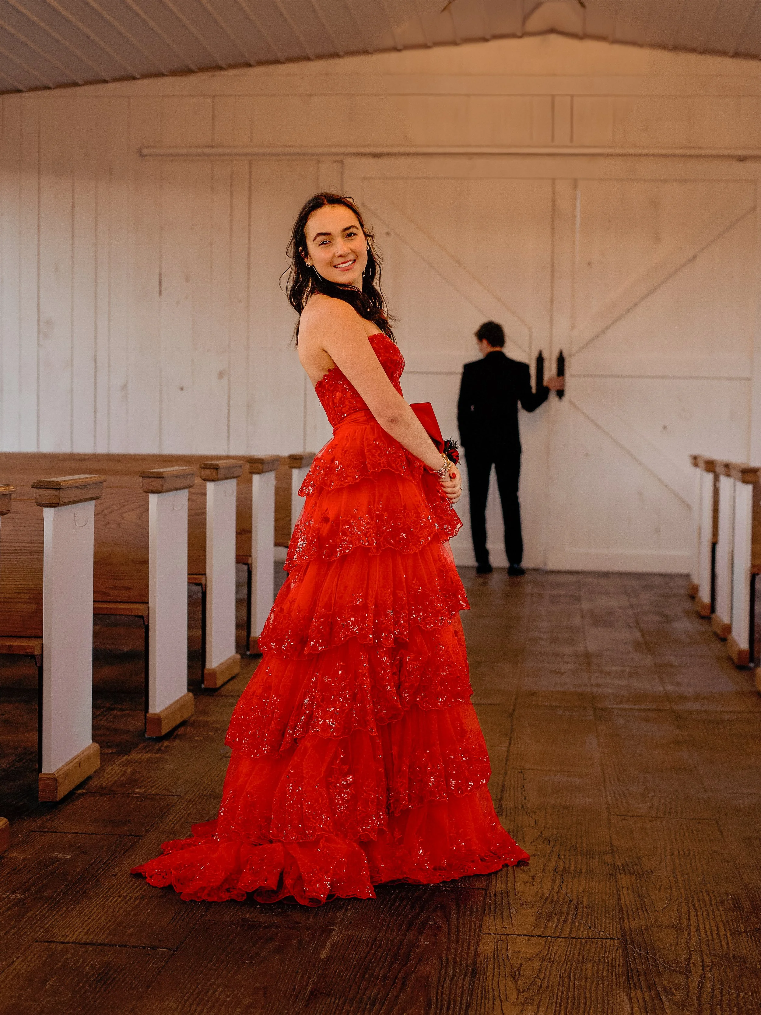 A young woman with dark hair smiling in a red strapless lace gown with a layered skirt at a rustic venue with wooden walls and floor, and a man in a black suit in the background.