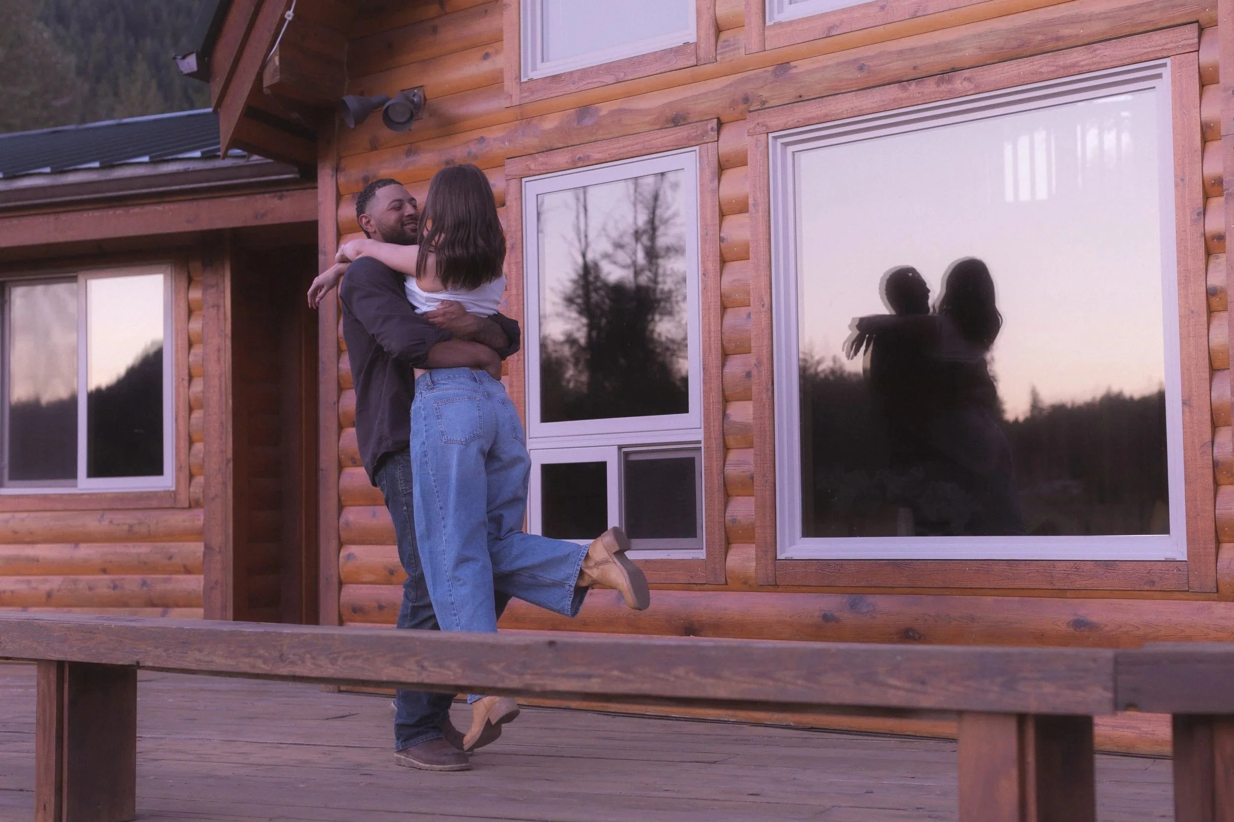 Man picking up woman and spinning her around. Reflection in window of cabin.