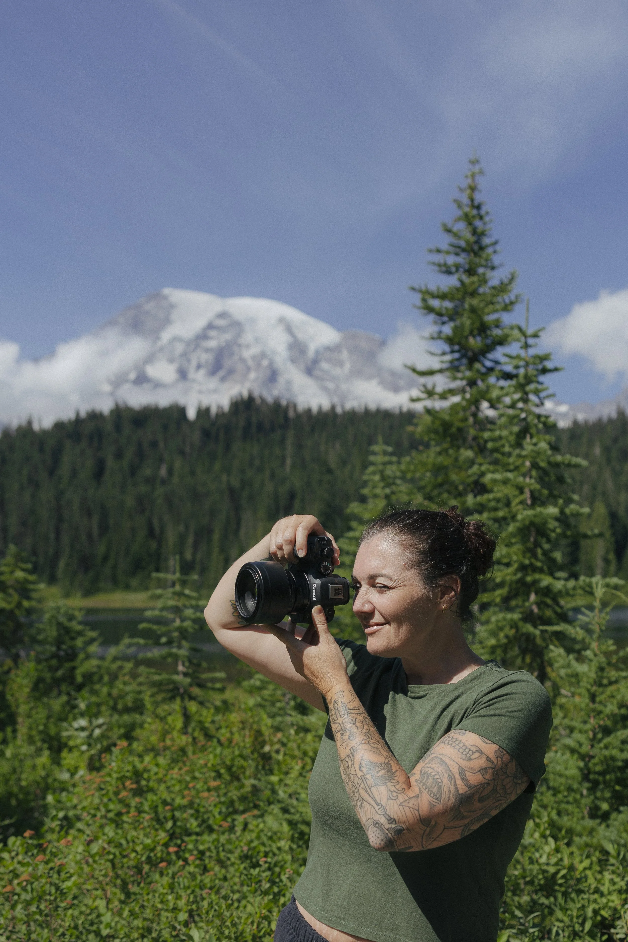 A woman with tattoos on her arms taking a picture with a camera outdoors. In the background, there is a mountain with snow on top, a forest of pine trees, and a partly cloudy sky.