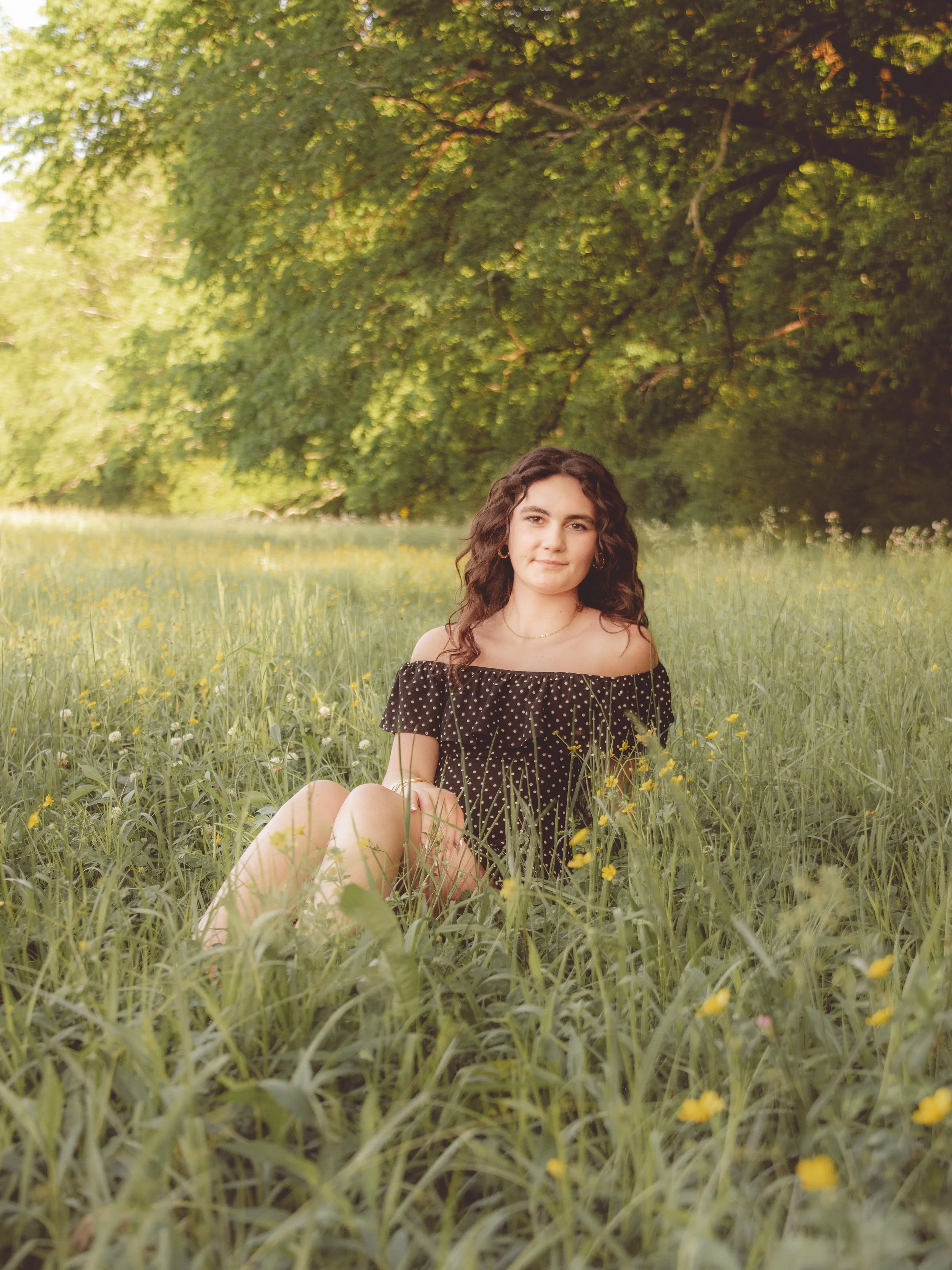 A young woman with curly brown hair sitting in a grassy field with yellow flowers, under a tree with green leaves, on a sunny day.