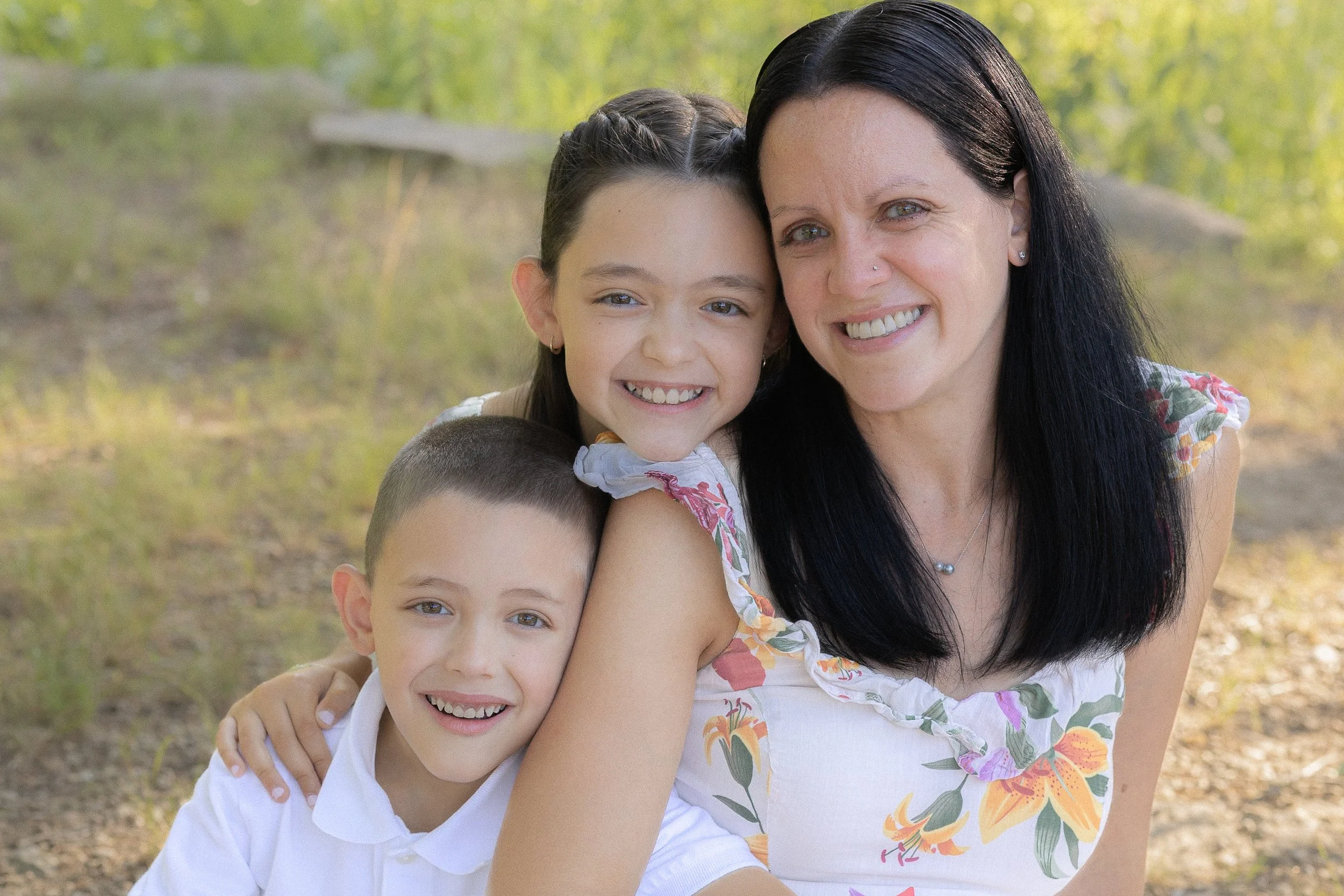 A woman with black hair and a nose piercing smiling while hugging two young boys outdoors in a wooded area, both boys smiling, in bright daylight.