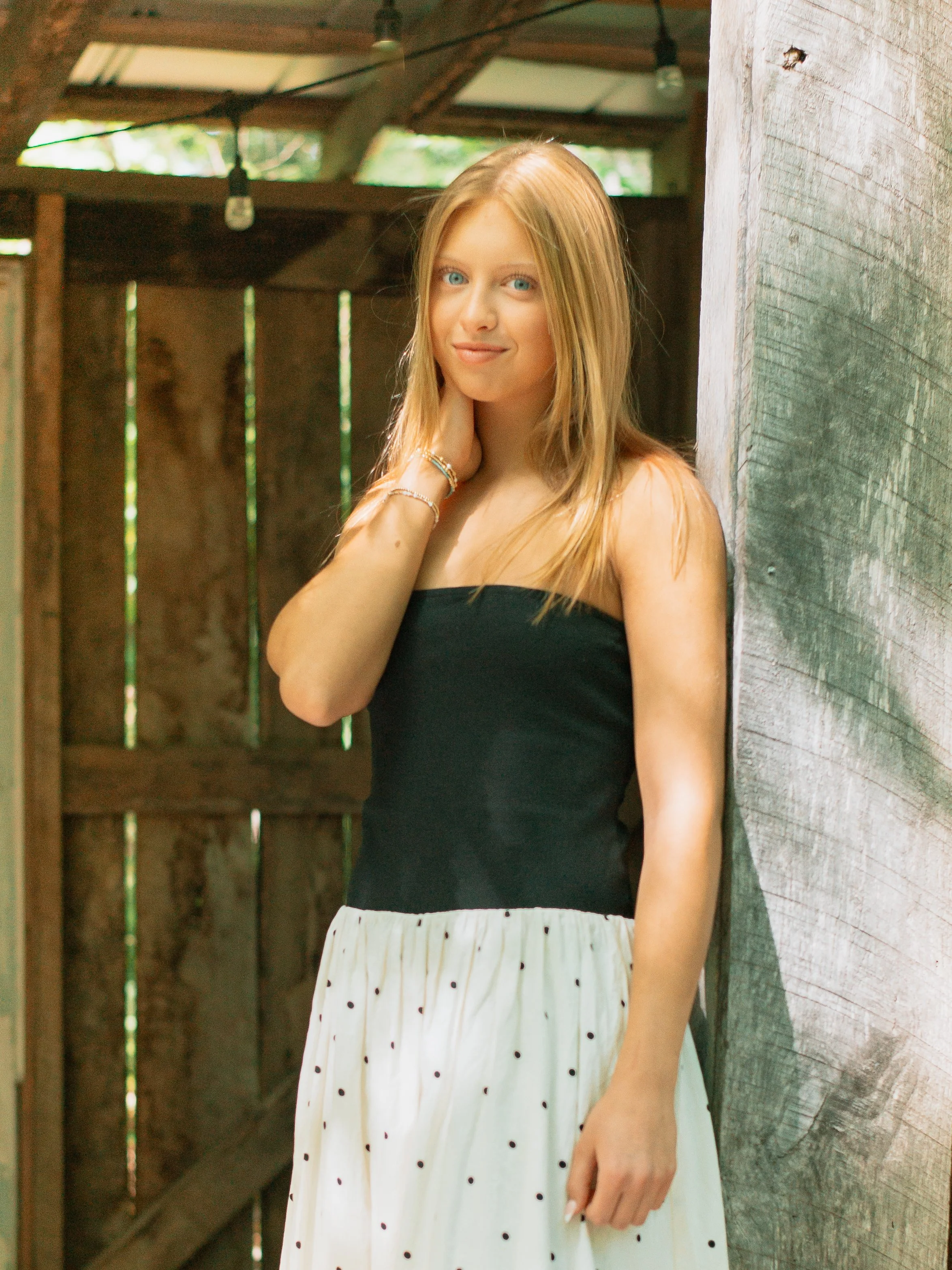 A young woman with long blonde hair and blue eyes, wearing a black strapless top and a white skirt with black polka dots, standing with her hand near her neck, smiling softly at the camera in a rustic wooden setting.