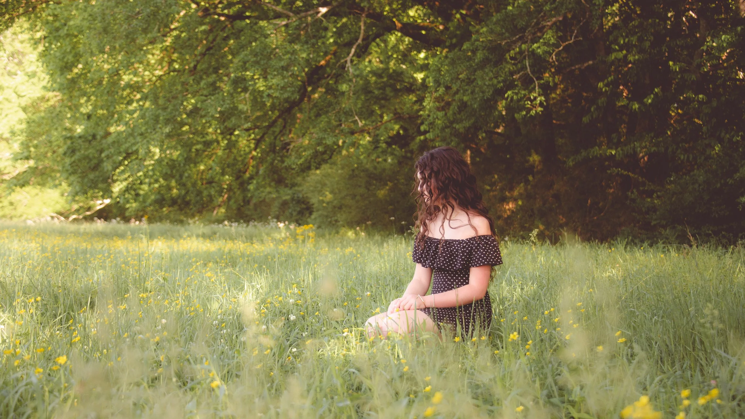 A woman with long curly hair wearing a black polka dot off-shoulder dress sitting in a field of yellow flowers near trees.
