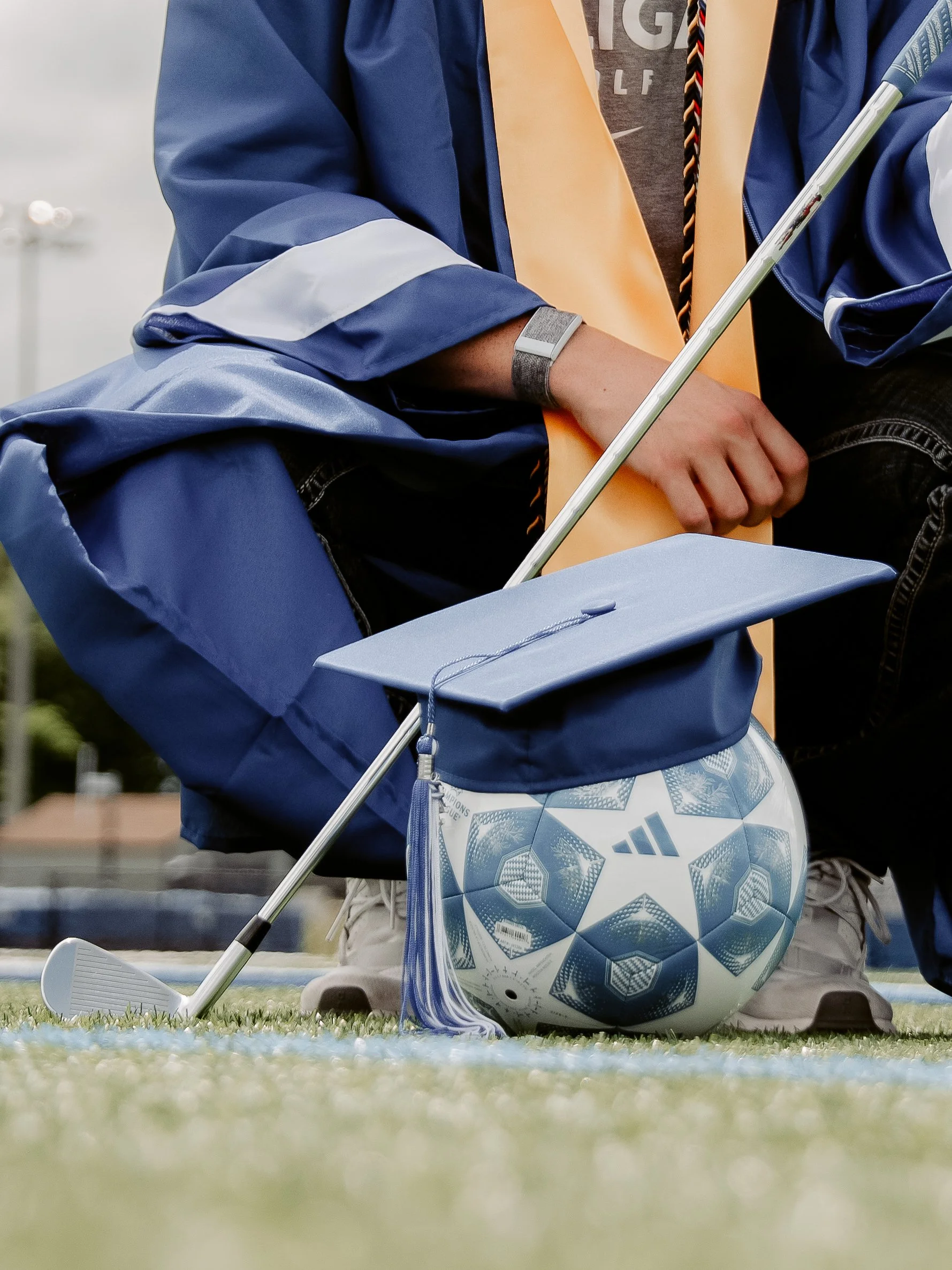 Close-up of a person kneeling on a football field with a golf club in their right hand, a blue sports bag, and a soccer ball nearby. The person is wearing a blue jacket and gray pants.
