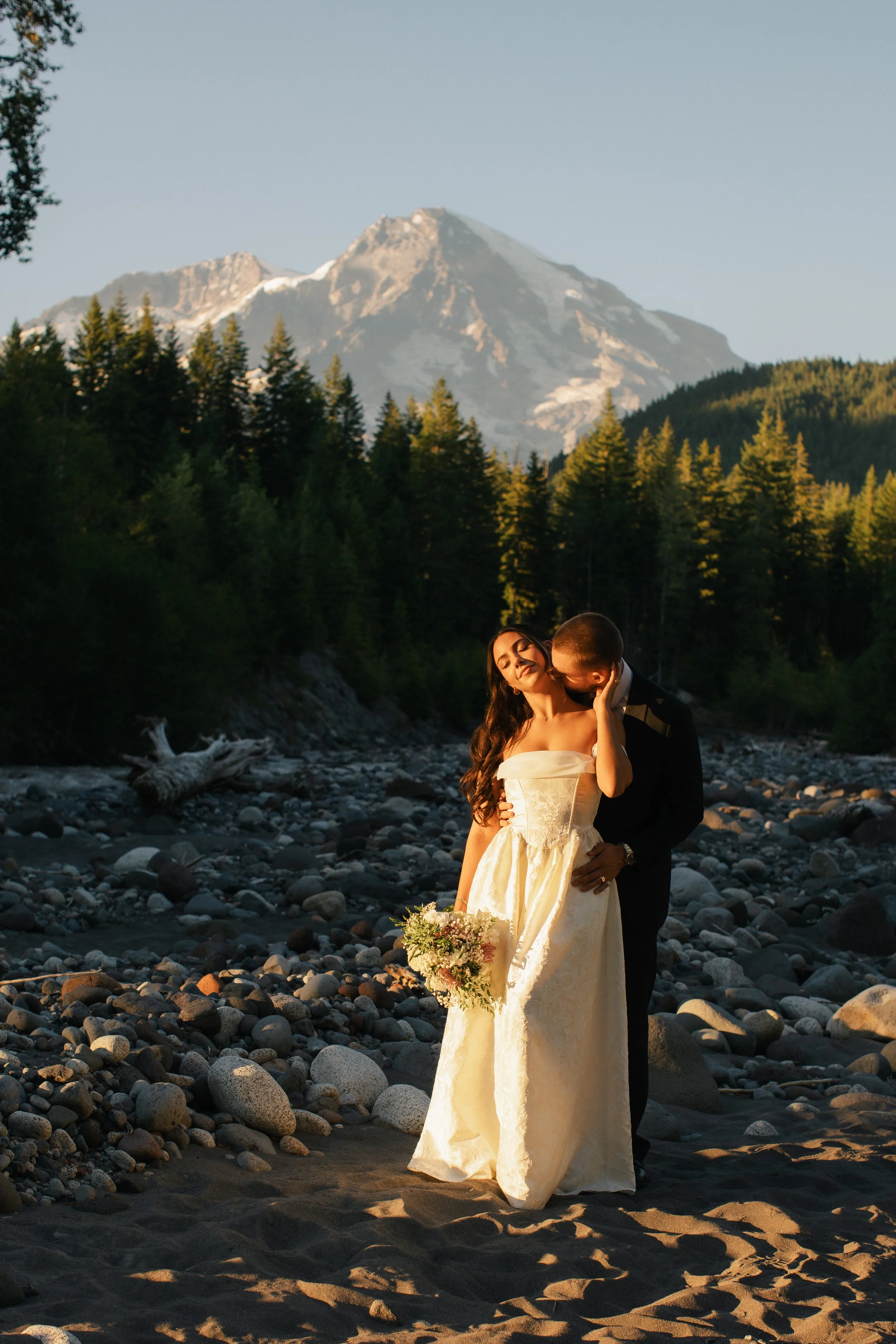 A bride and groom stand on a rocky beach with a mountain in the background. The bride is smiling and holding a bouquet, while the groom is embracing her from behind, kissing her cheek. The scene is illuminated by warm sunlight.