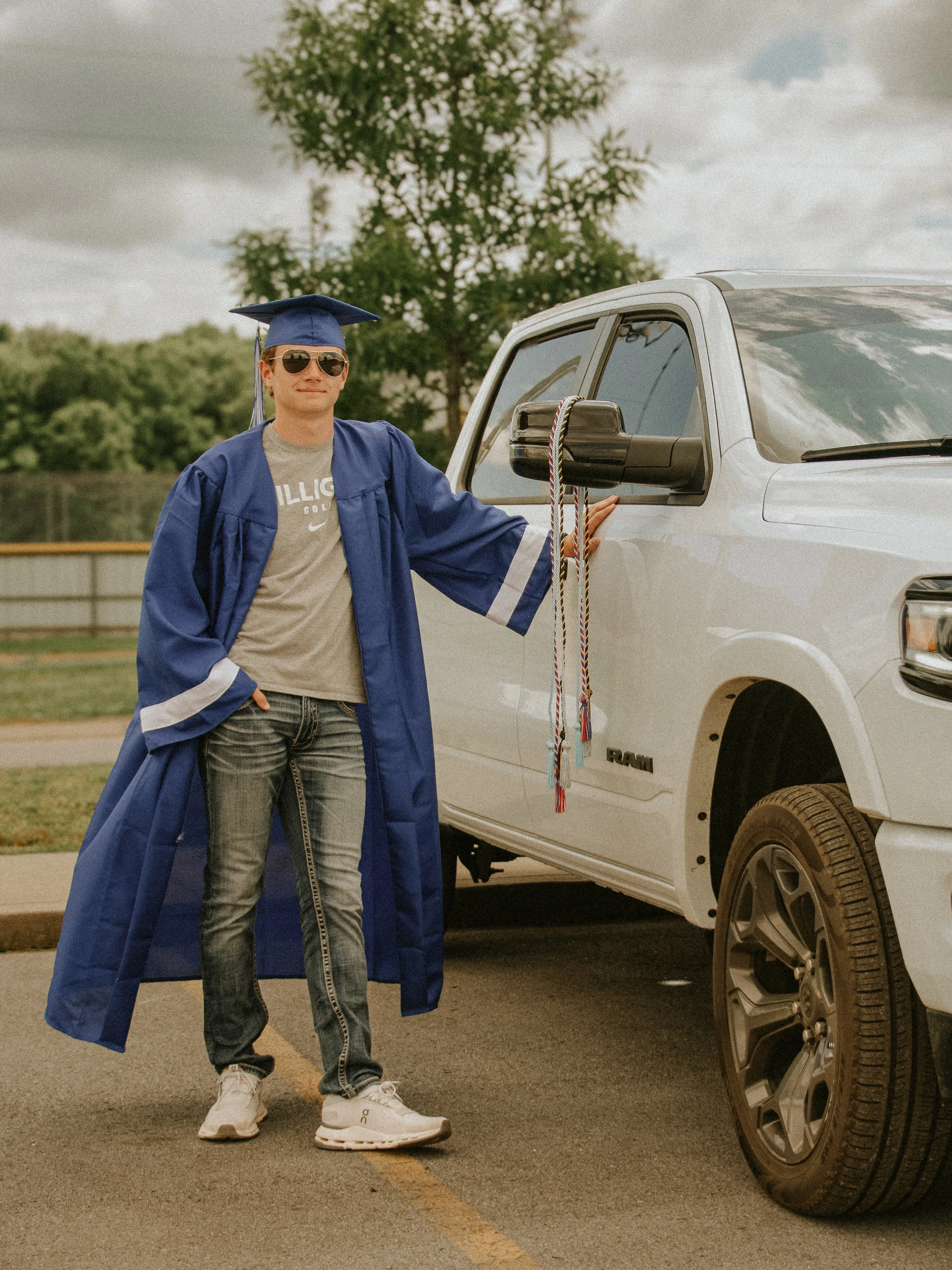 A young man wearing a blue graduation cap and gown, sunglasses, a gray t-shirt, jeans, and white sneakers standing next to a white RAM pickup truck outdoors. He has one hand on the truck and is smiling at the camera.