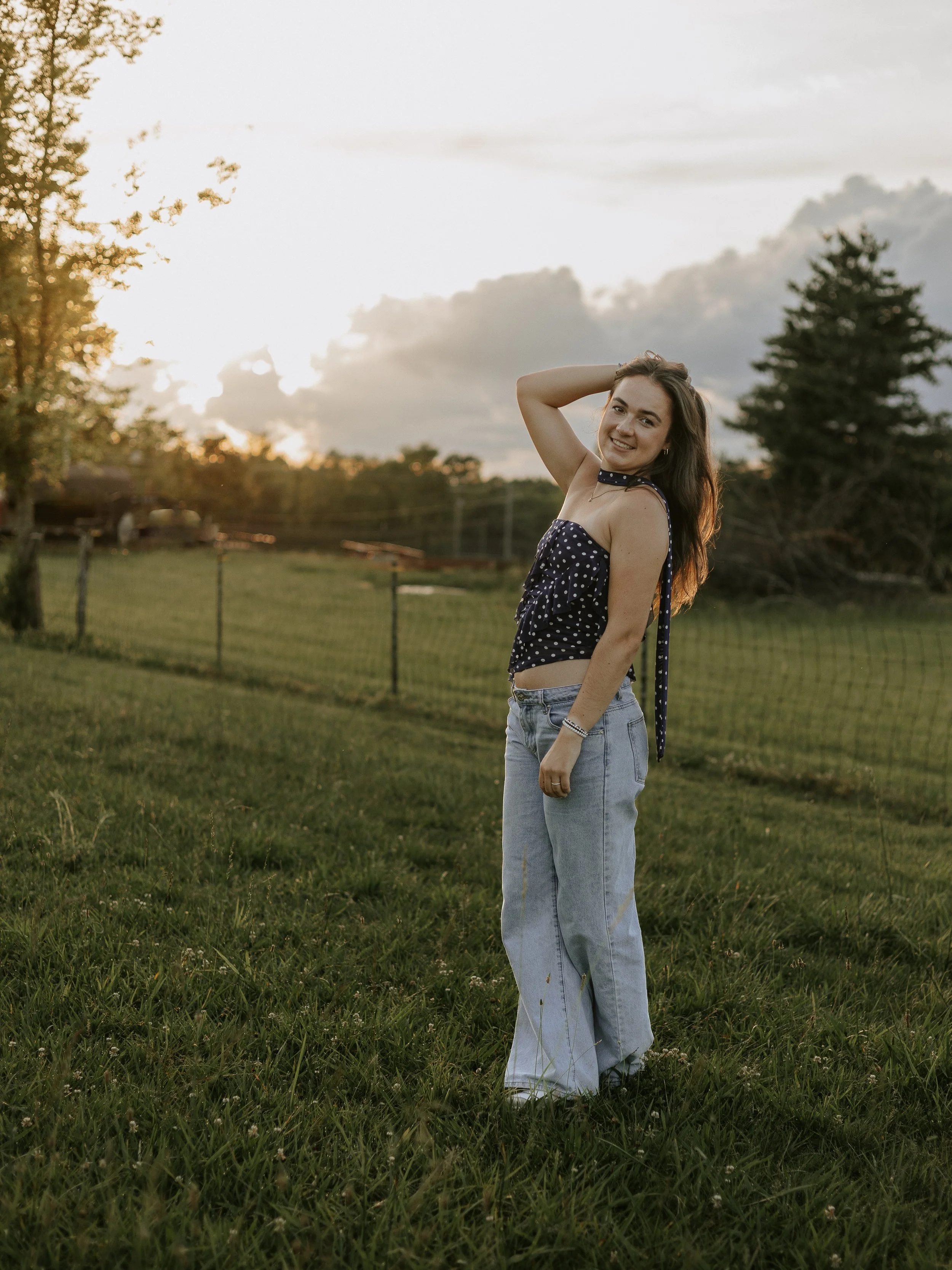 A young woman with long brown hair, wearing a navy blue polka-dot crop top and light blue jeans, standing outdoors on a grassy field with trees and a fence in the background. The sky is partly cloudy and the sun is setting, casting warm light.