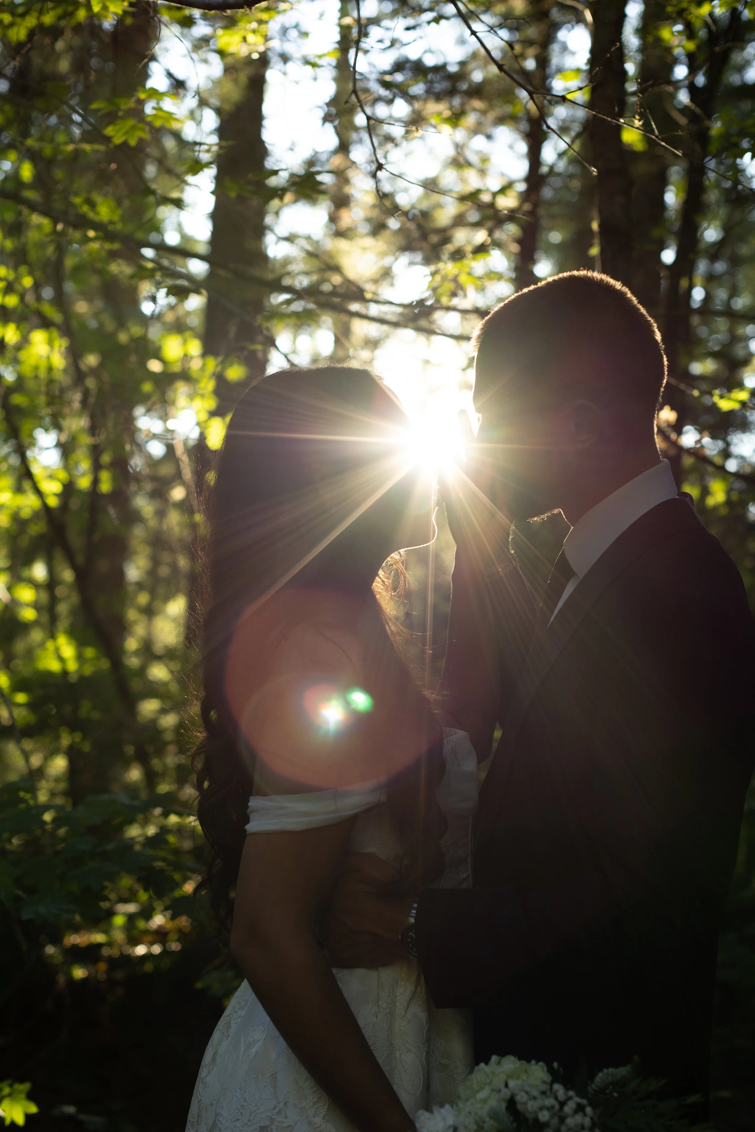 Silhouette of a bride and groom standing closely in a wooded area, with sunlight shining between them and trees in the background.