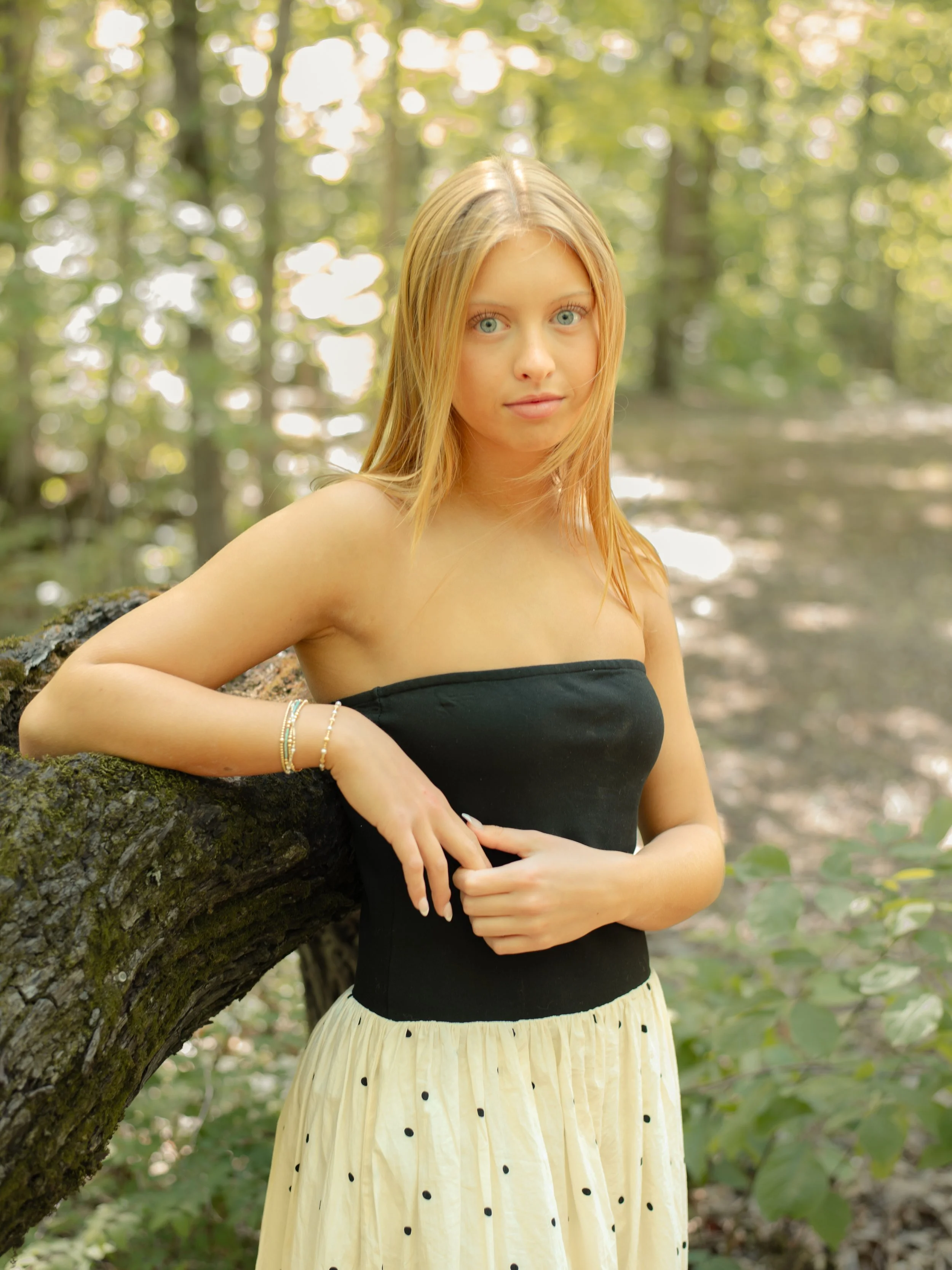 A young woman with long blonde hair and blue eyes leaning against a tree in a forest, wearing a black strapless top and a yellow skirt with black polka dots, adorned with bracelets on her wrist.