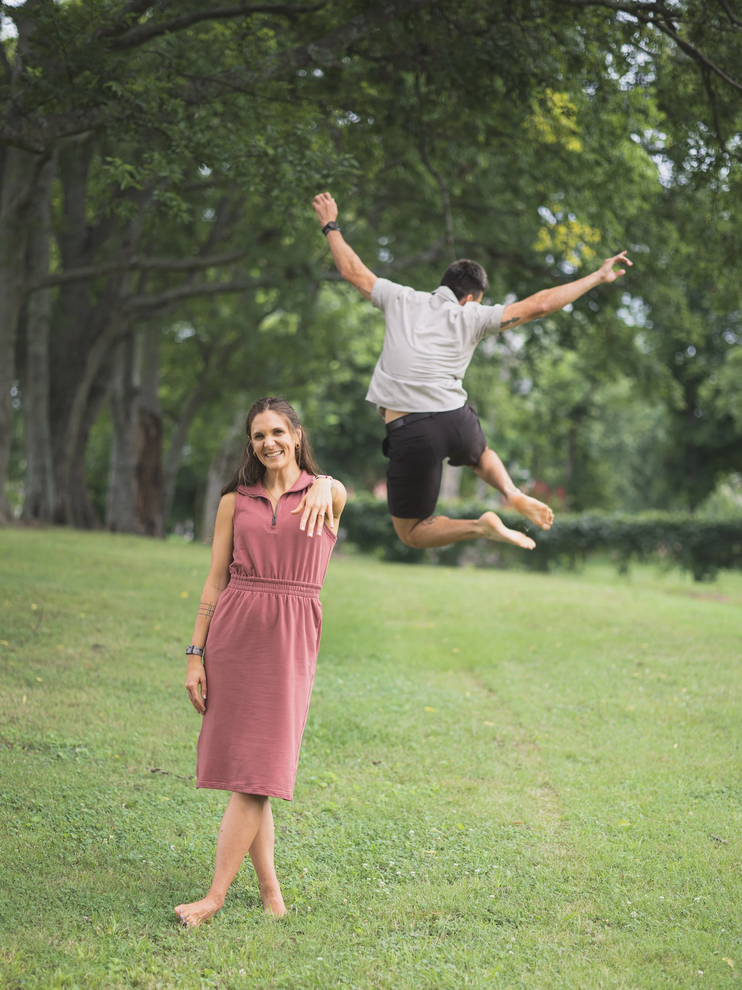 A woman in a maroon dress standing on grass with a man jumping in the air behind her in a park after their engagement.