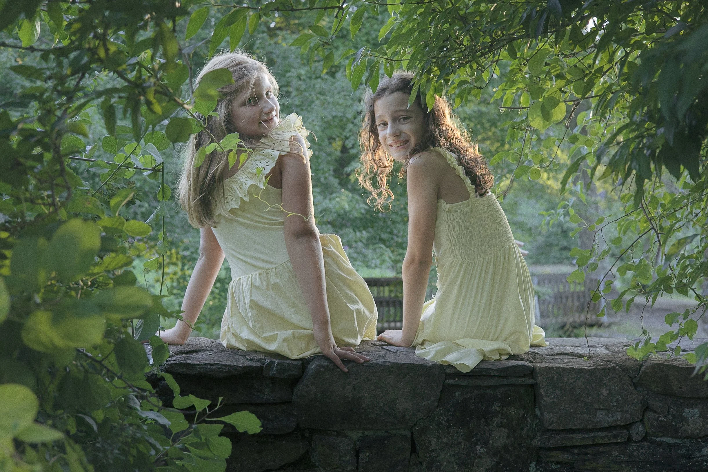 Two little girls in yellow dresses sitting on a rock wall with green foliage around them