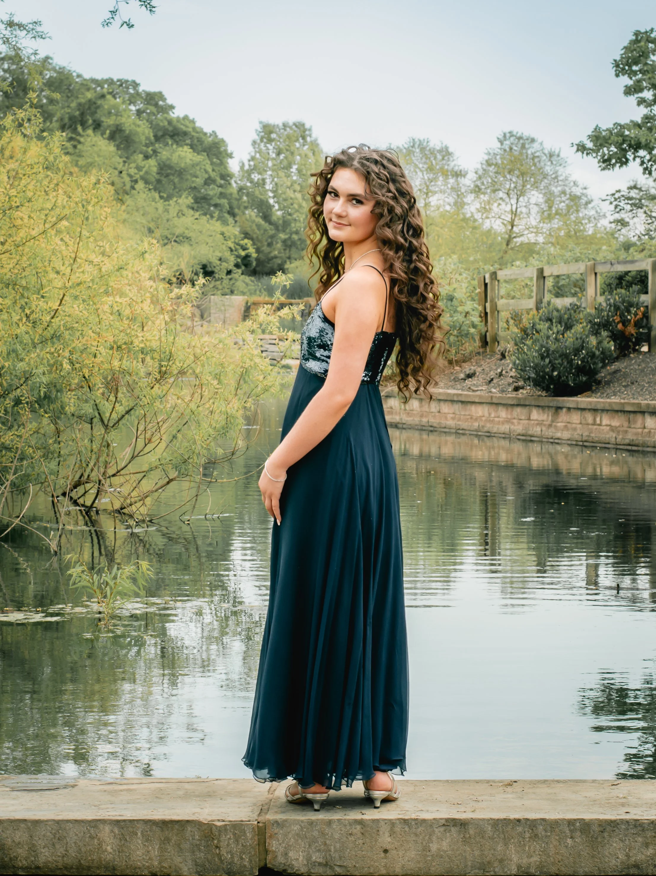 A young woman with long curly hair in an elegant blue dress standing on a stone dock by a lake, surrounded by trees and greenery.