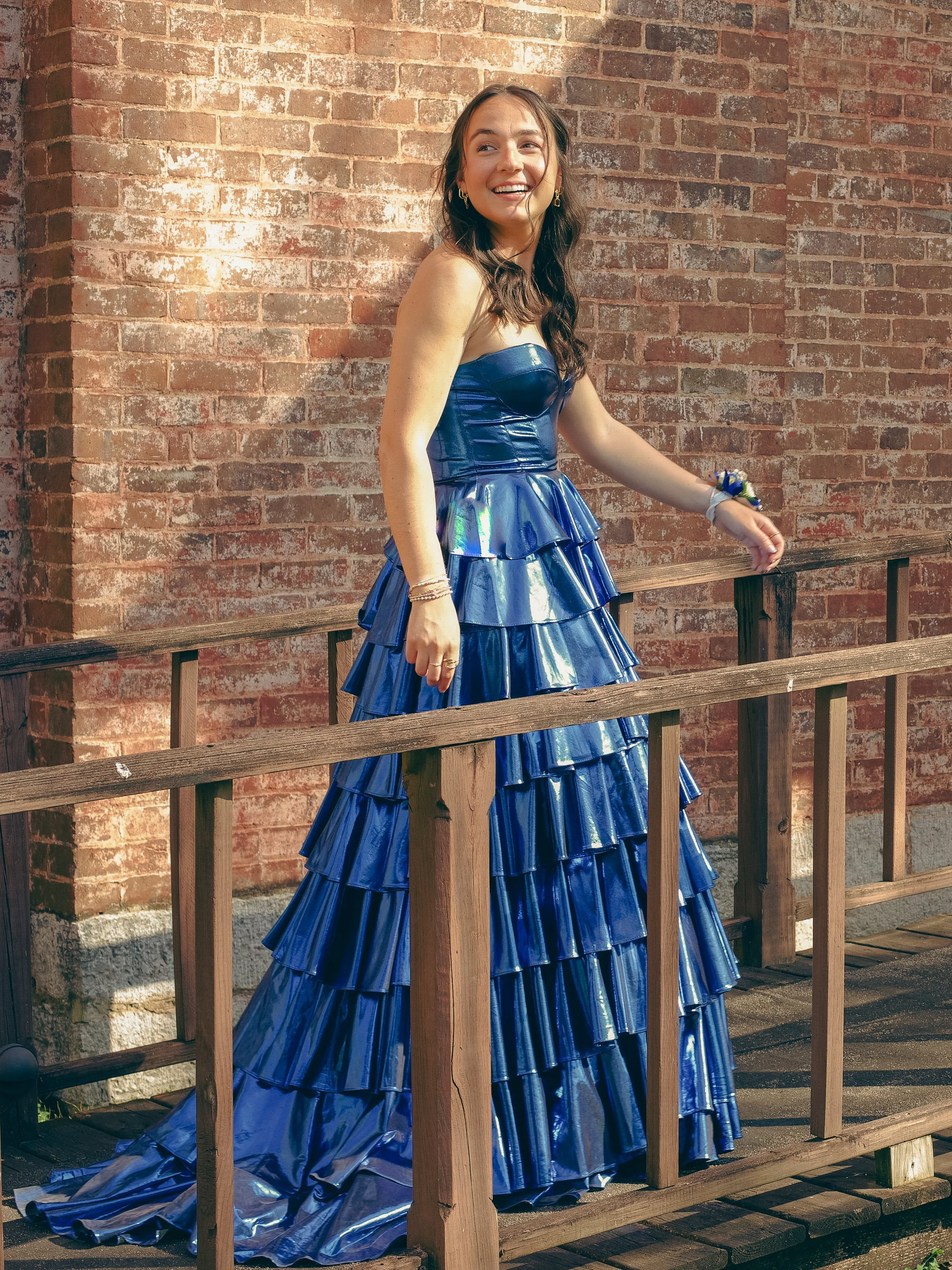 A young woman with long dark hair, wearing a shiny blue strapless gown with tiered ruffles, is standing outdoors next to a brick wall and a wooden railing, smiling and looking off to the side.