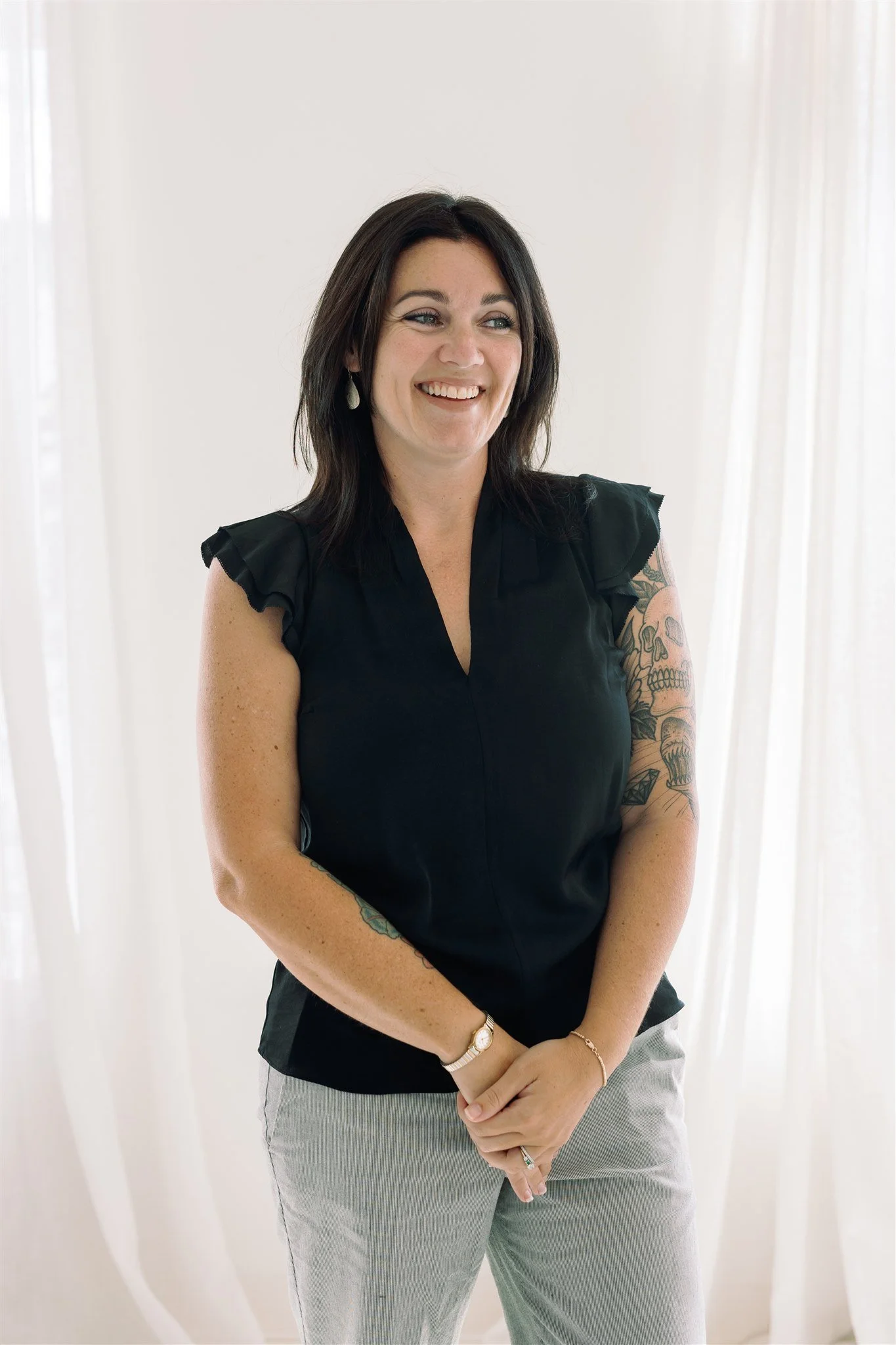 A smiling woman with dark hair, wearing a black sleeveless top with ruffled shoulders, gray pants, and jewelry, standing in front of white curtains.