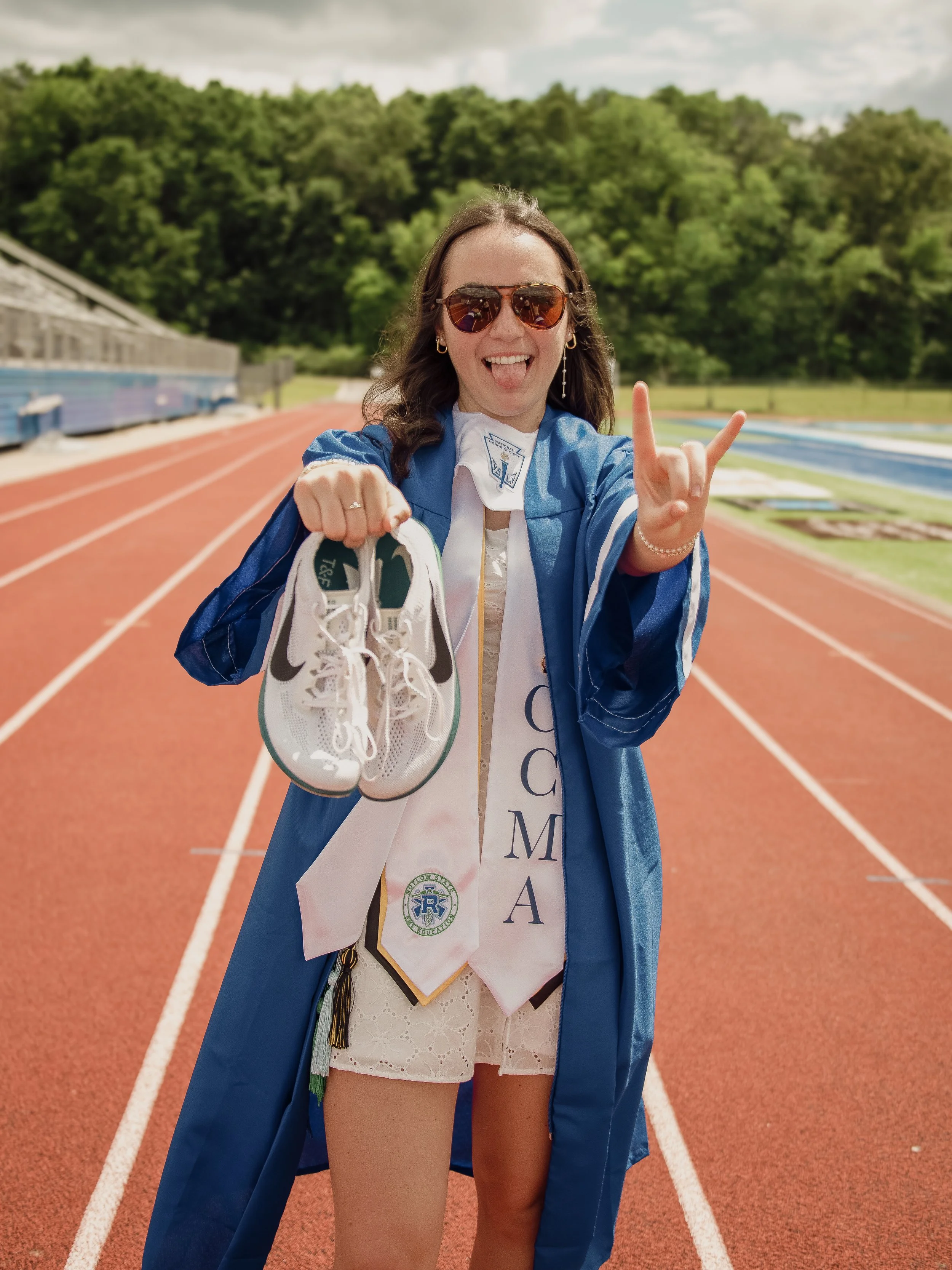 A joyful woman wearing sunglasses and a graduation gown standing on a running track, holding her shoes, with a medal and sash, in an outdoor stadium with green trees in the background.