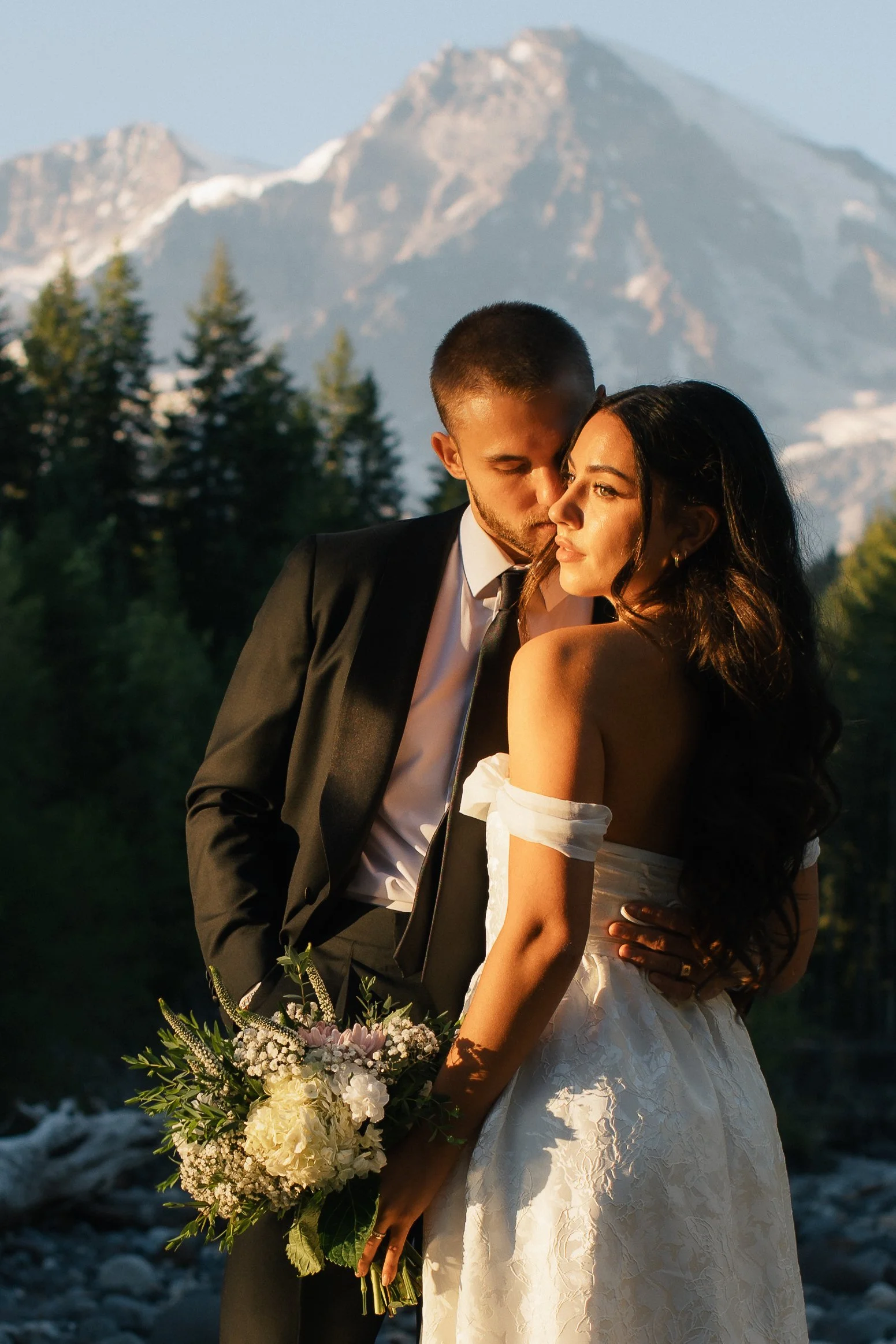 A newlywed couple standing outdoors with mountain scenery in the background, the groom with short dark hair and in a black tuxedo and the bride in a white strapless wedding gown holding a bouquet.