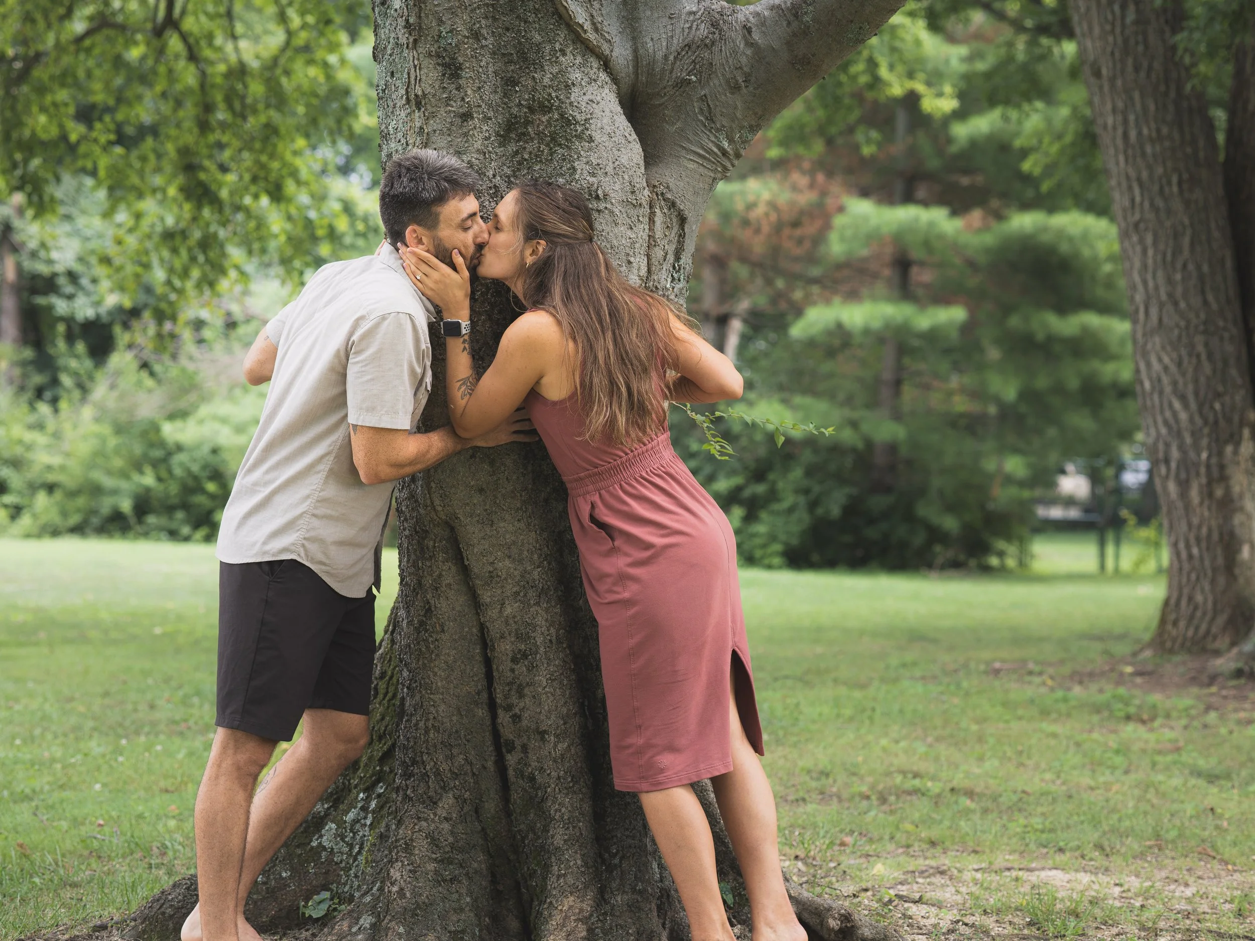 Man and woman kissing around a giant tree trunk after getting engaged in a park