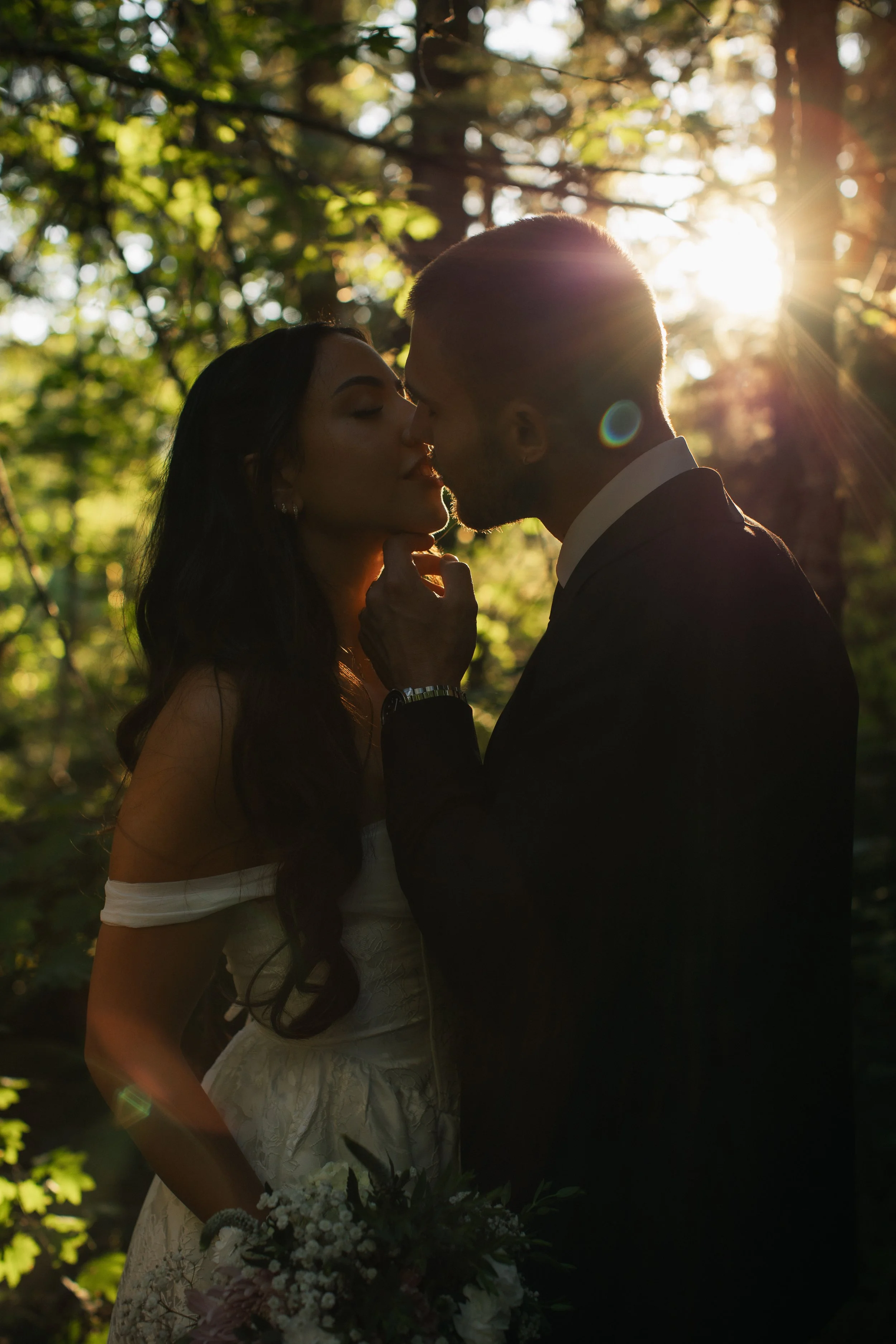 A couple sharing a kiss outdoors during sunset, with sunlight filtering through trees in the background, and the woman holding a bouquet of flowers.