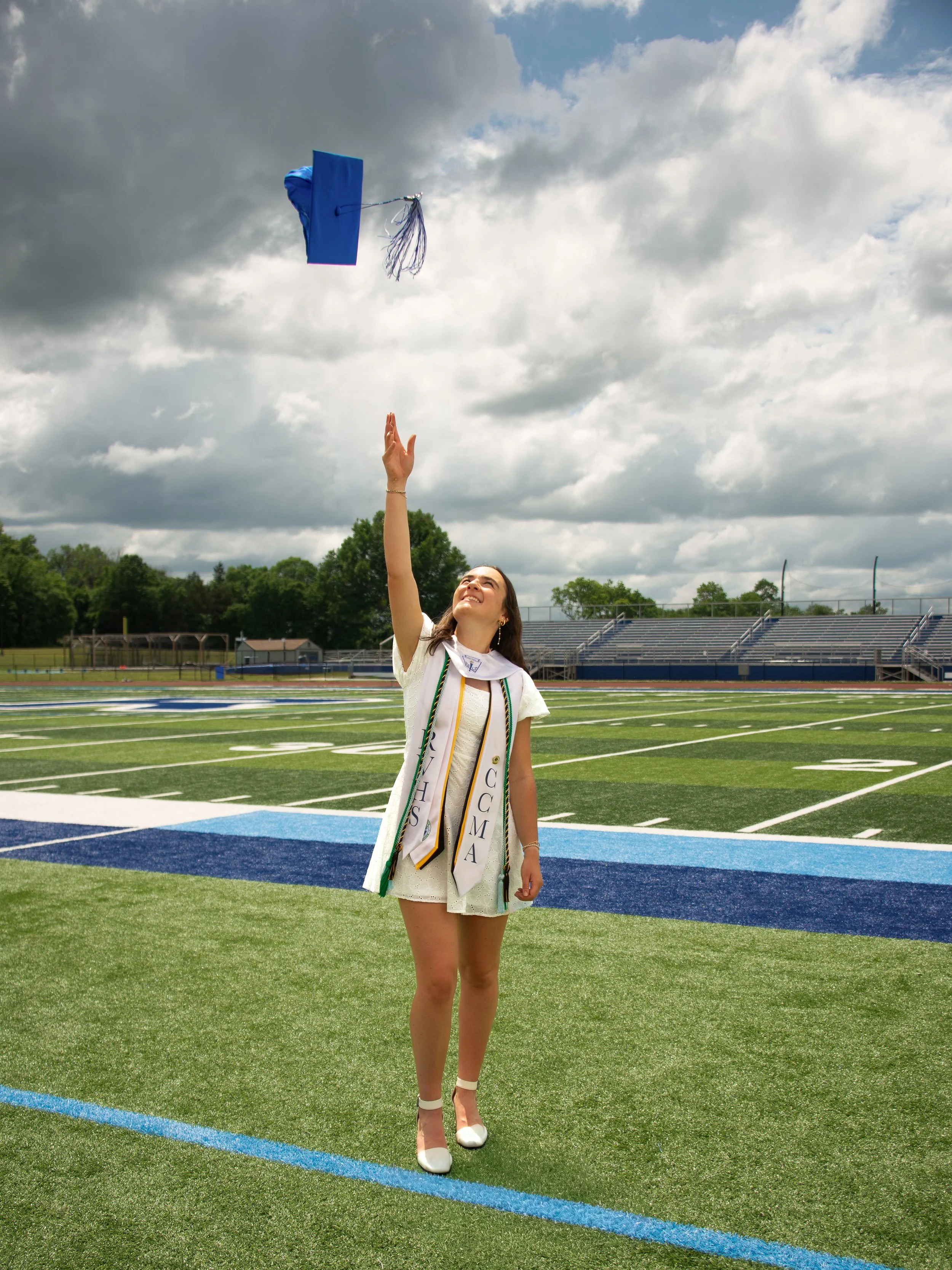 A young woman wearing a graduation cap and gown with medals and cords, standing on a football field, tossing her cap into the air, with cloudy skies and stadium bleachers in the background.