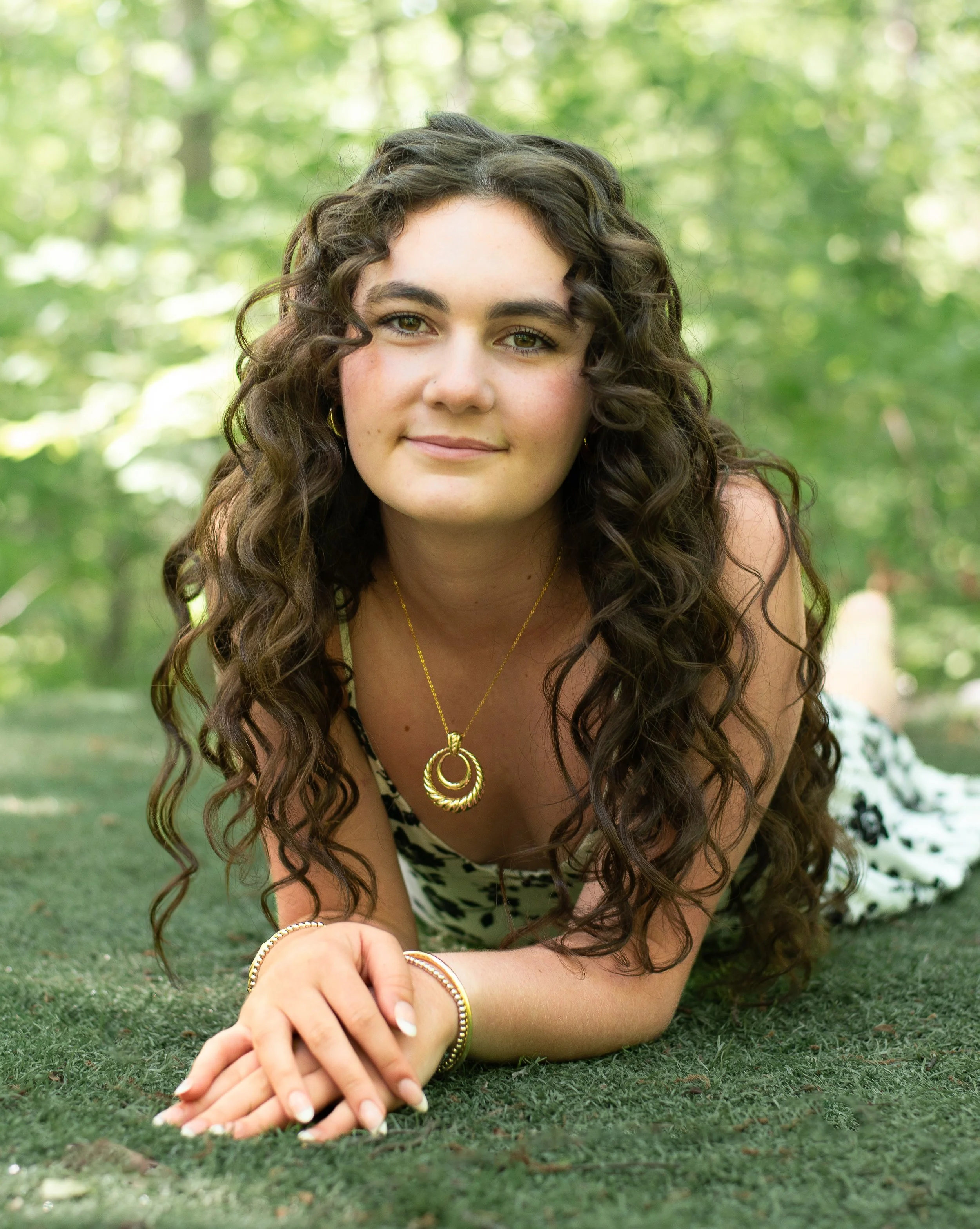 A young woman with curly brown hair and light skin lying on a grassy surface outdoors with a blurred green foliage background, smiling softly at the camera, wearing gold jewelry including a necklace, earrings, bracelet, and rings.