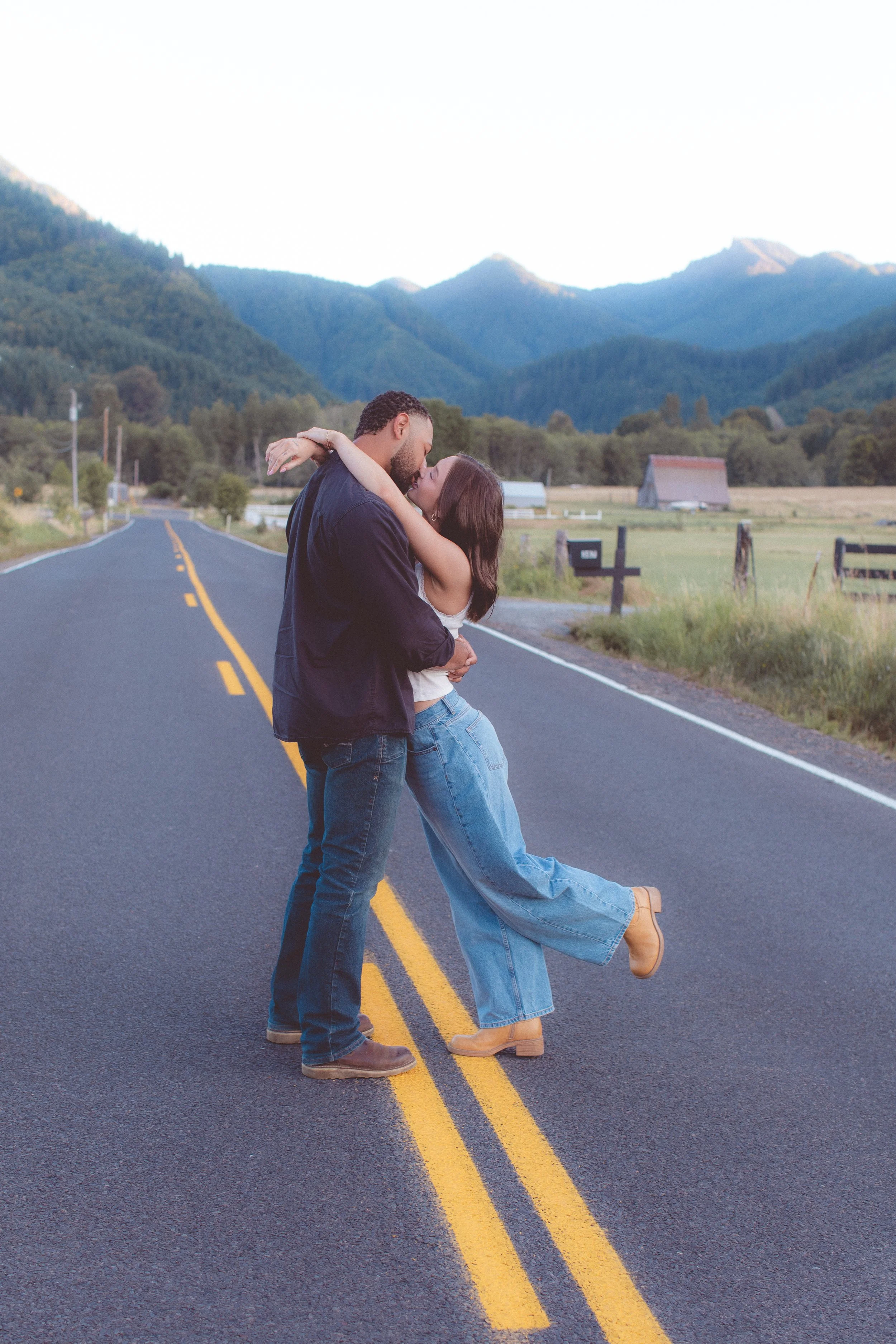 Man and woman kissing in the road with mountains in the background. 