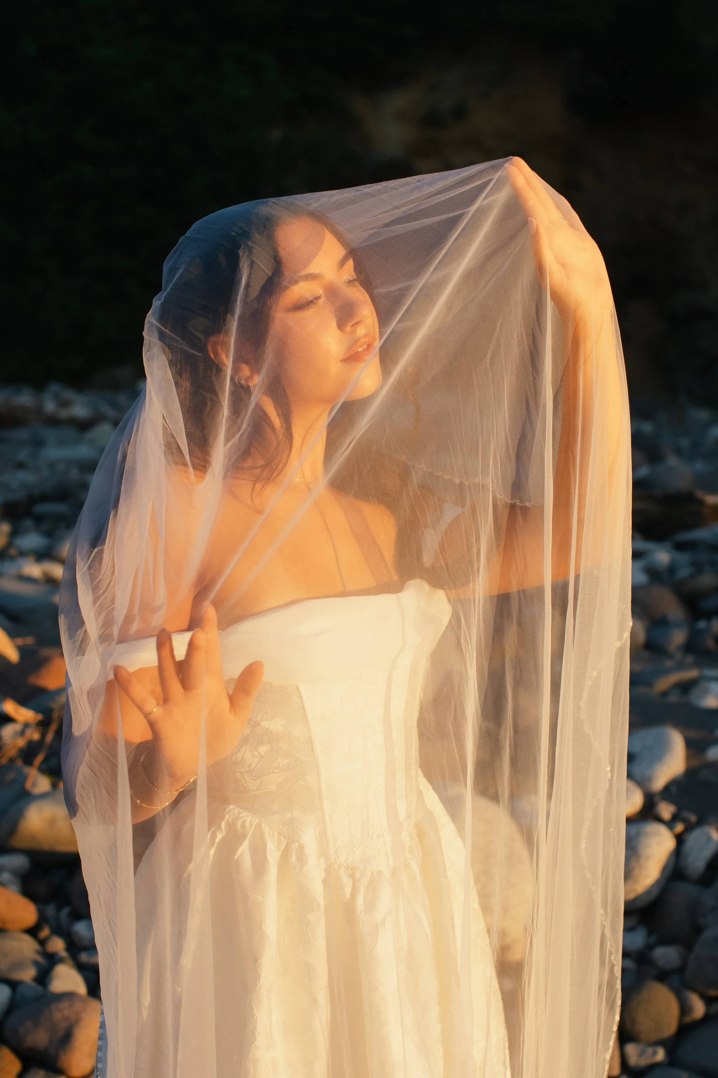 A woman in a white dress with a sheer veil, standing outdoors on rocks, her eyes closed and face serene in sunlight.