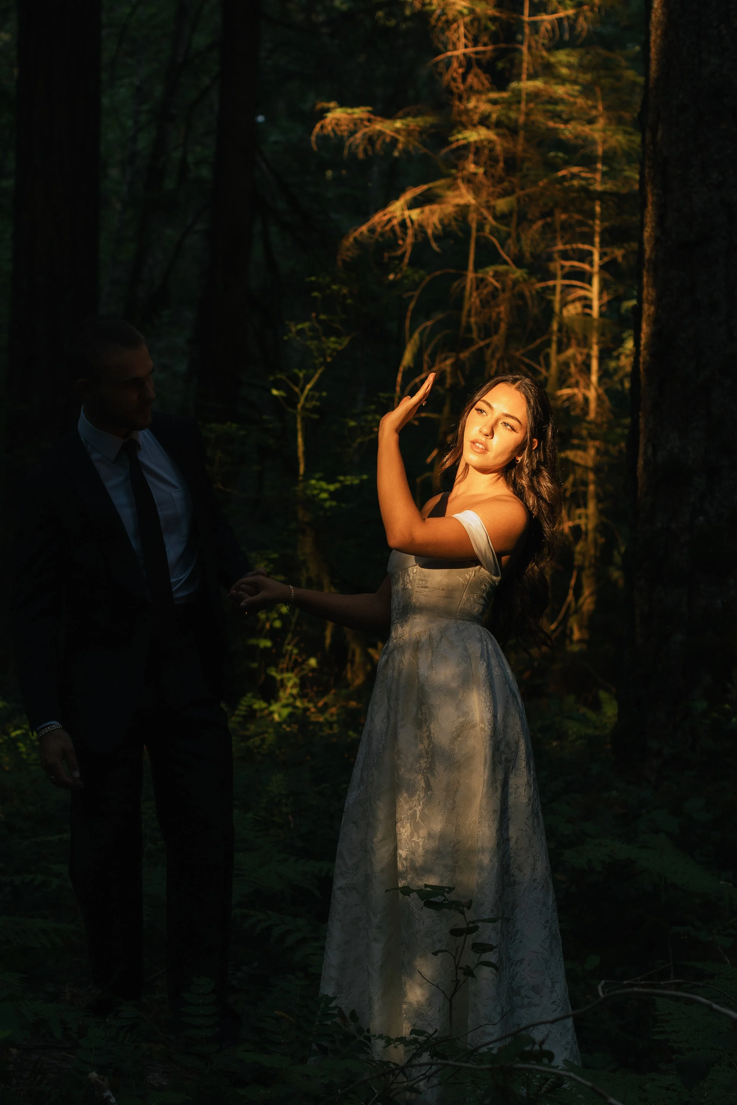 A woman in a white lace wedding dress is standing in a forest at dusk, illuminated by a warm, soft light. A man in a suit is holding her hand, standing nearby, partially shadowed. The background features tall trees with dark foliage.