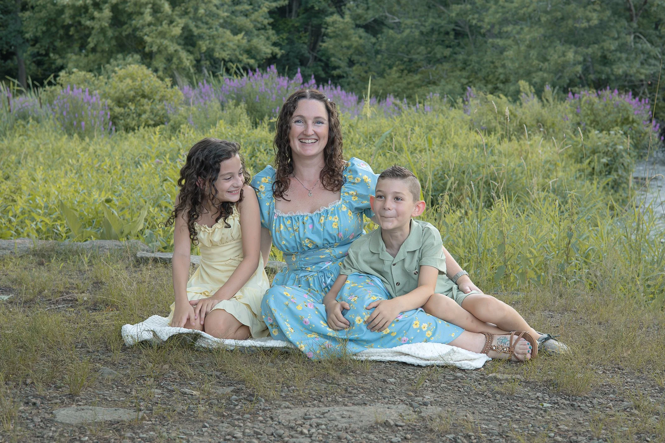A woman with three children sitting on a blanket outdoors near a river, with green trees and purple flowers in the background, smiling and enjoying a nature outing.