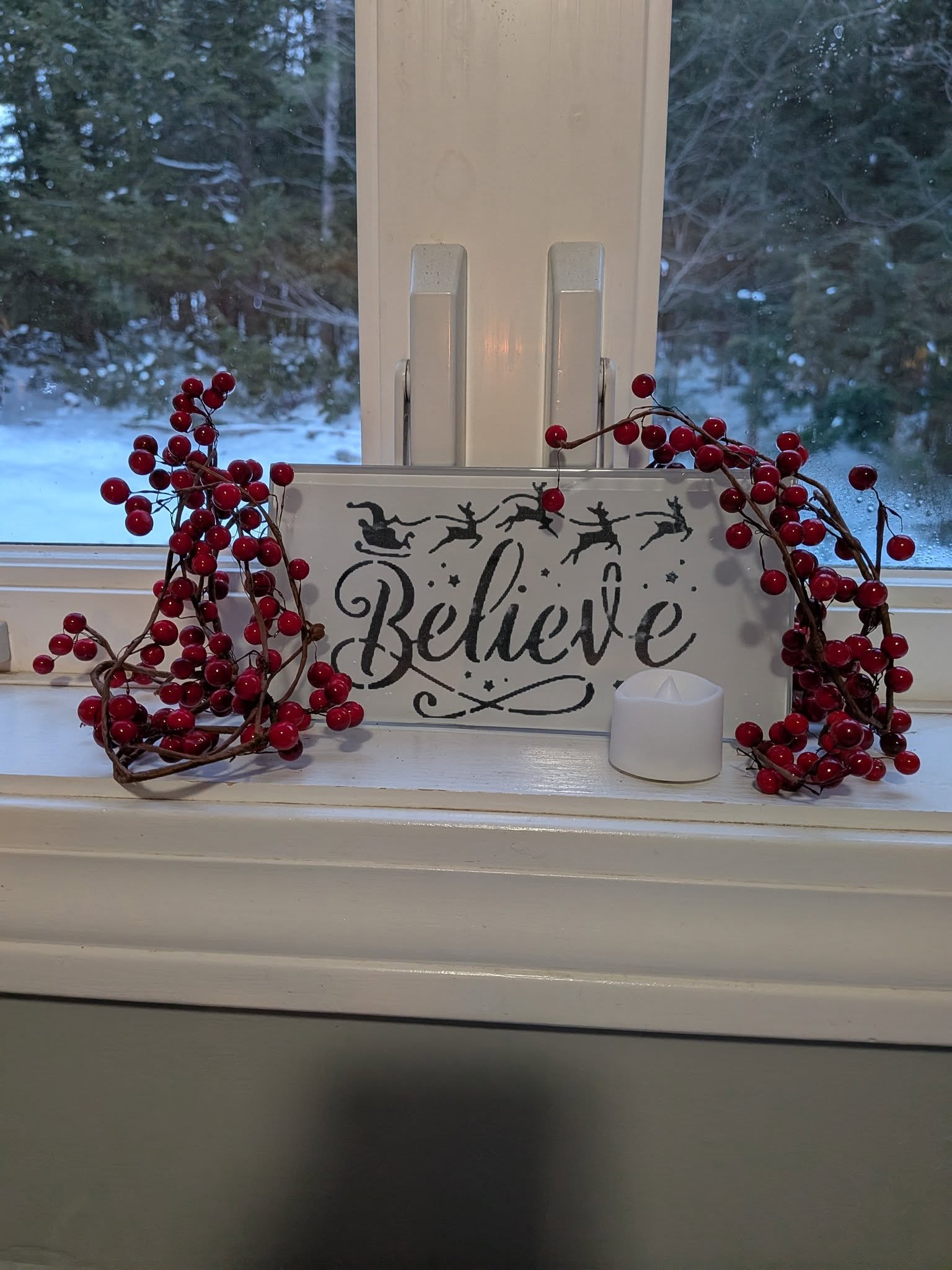 A windowsill with decorative items, including a small sign with the word 'Believe' and reindeer silhouettes, red berry garland, a small white candle, and a view of snow-covered trees outside.