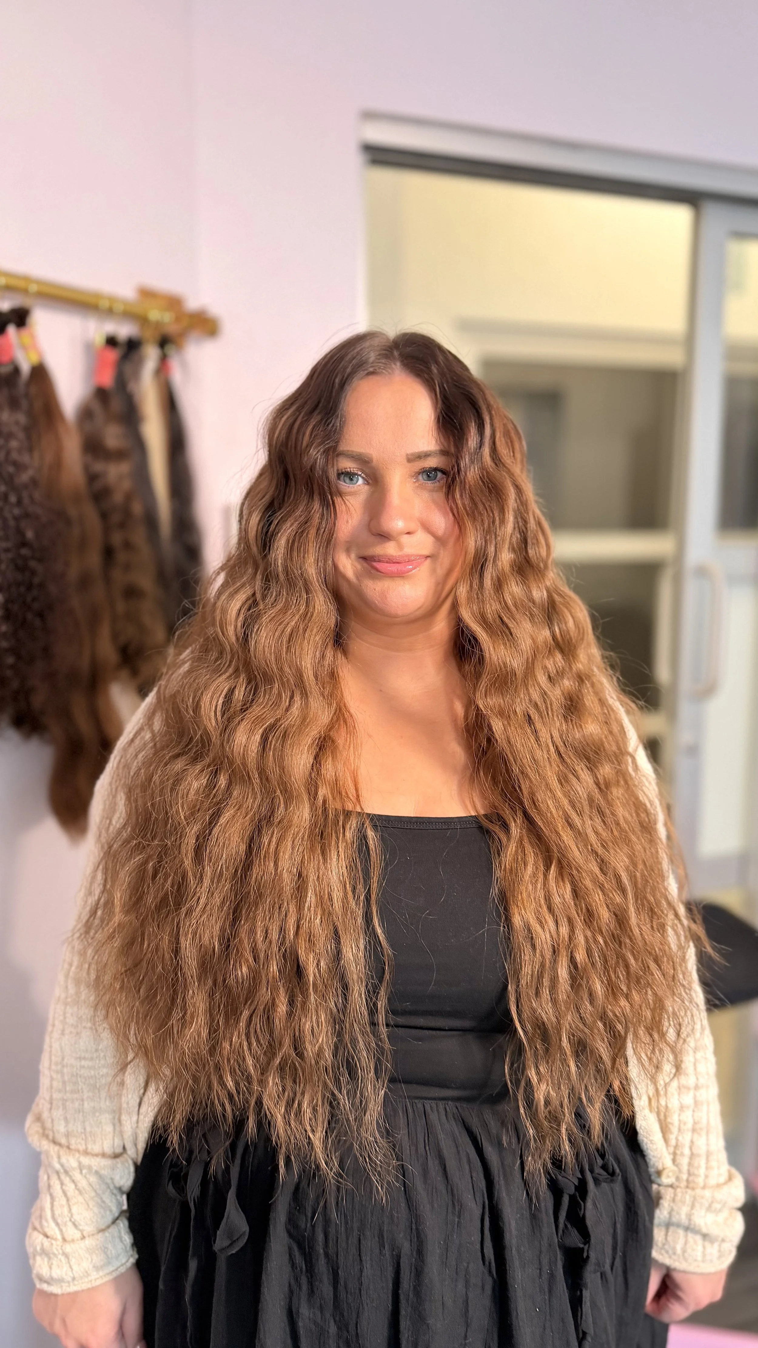 A woman with tanned skin and long, wavy, reddish-brown hair standing indoors near a wall with hair extensions hanging on a rack behind her. She is wearing a black top and a beige cardigan, smiling softly at the camera.
