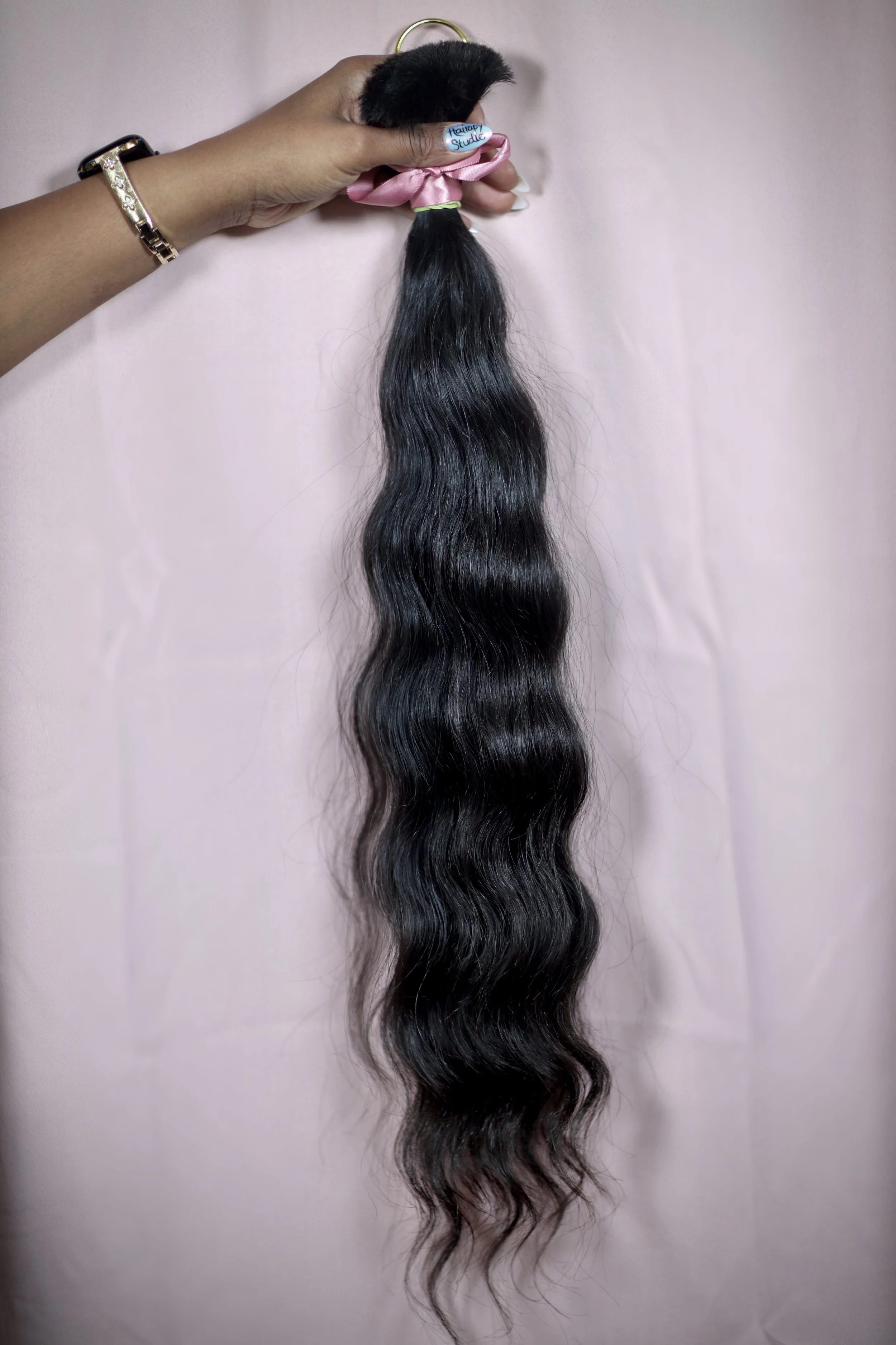 A hand holding a wavy, black hair extension with a pink ribbon near the top, against a light pink background.