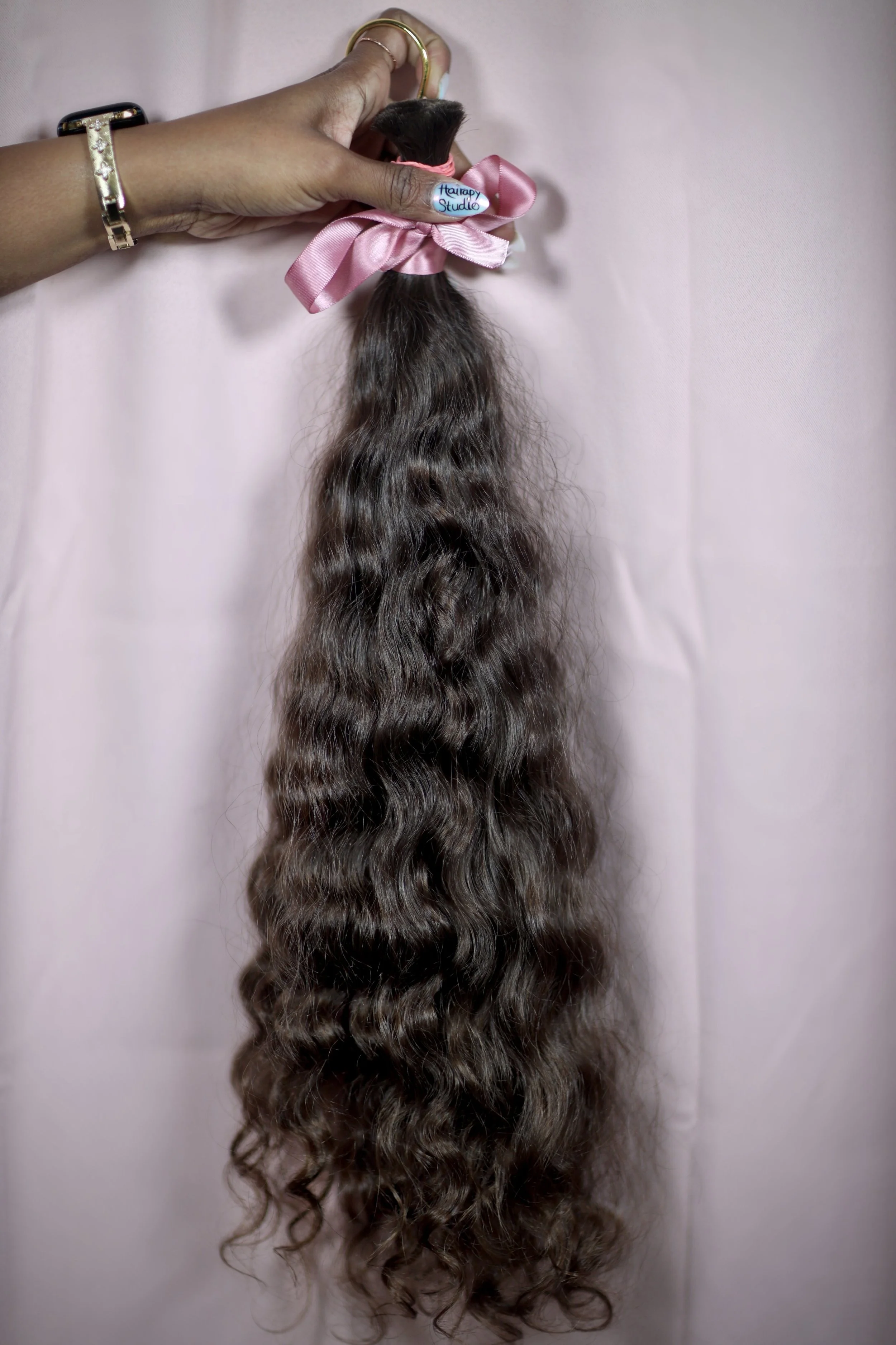 A hand holding a bundle of long, wavy, dark brown hair, tied with a pink ribbon, against a light pink background.